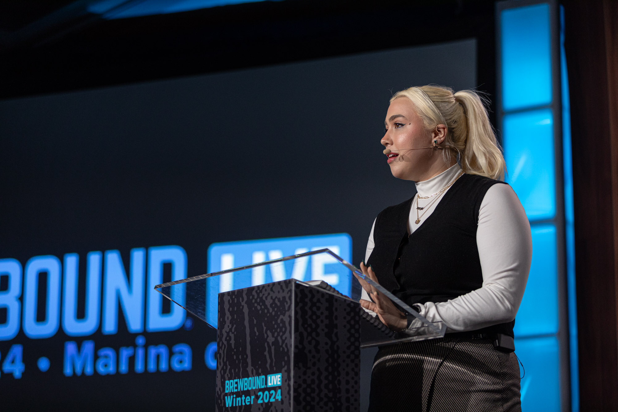 A blonde woman in a black vest and white top speaks from a podium on stage.