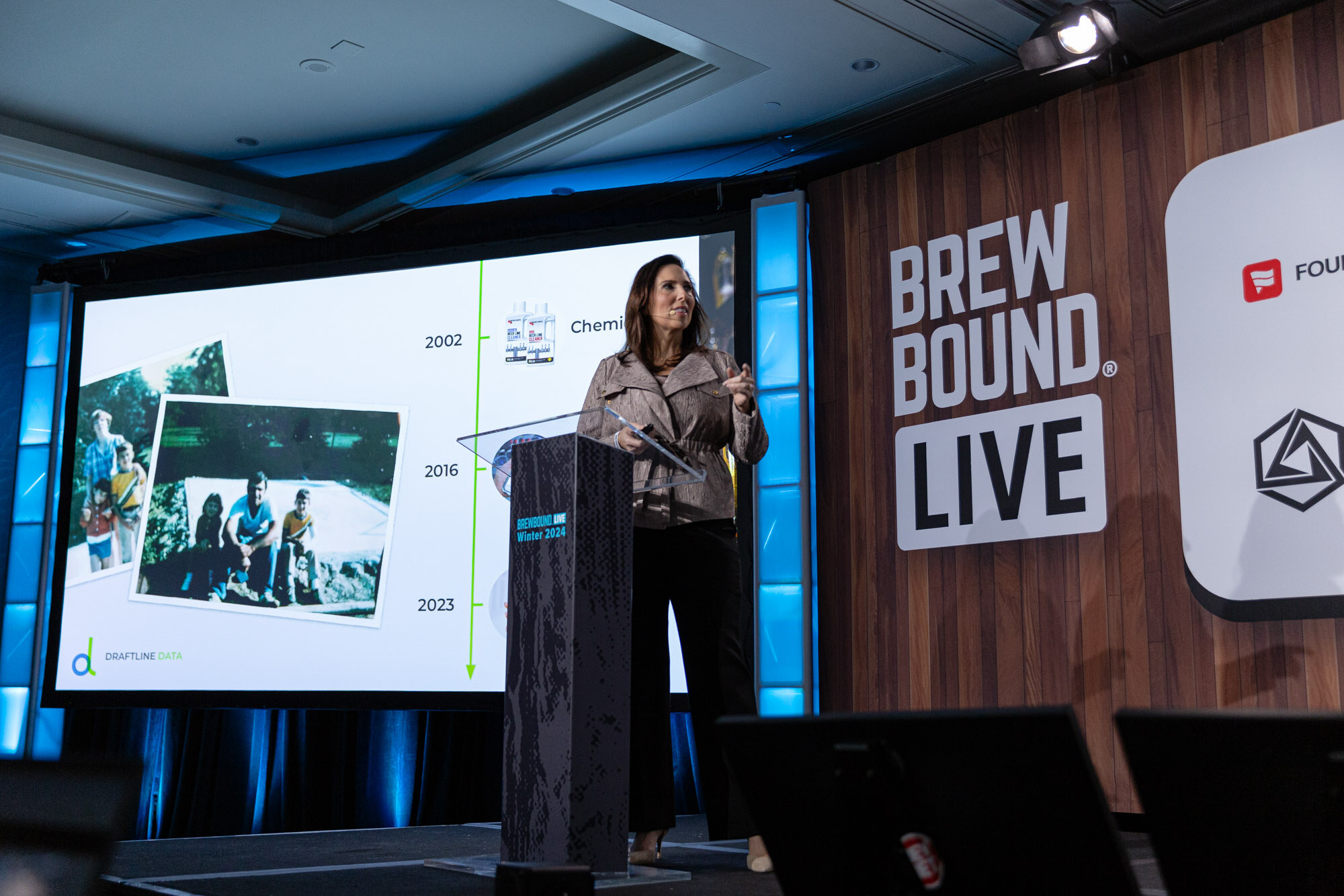 A woman stands at a podium next to a screen showing a timeline with old photographs from 2002, 2016 and 2023.