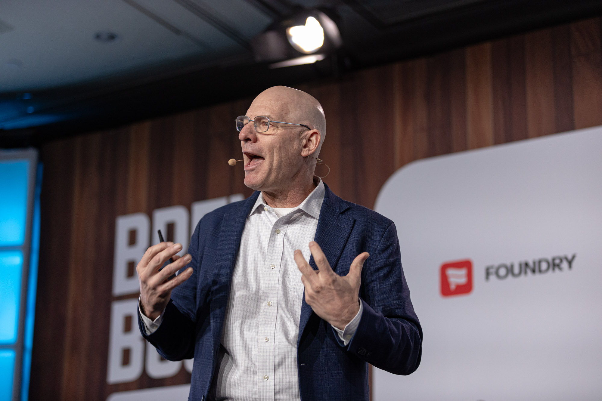 A bald man with glasses and a navy blazer gestures with both hands while speaking on stage.