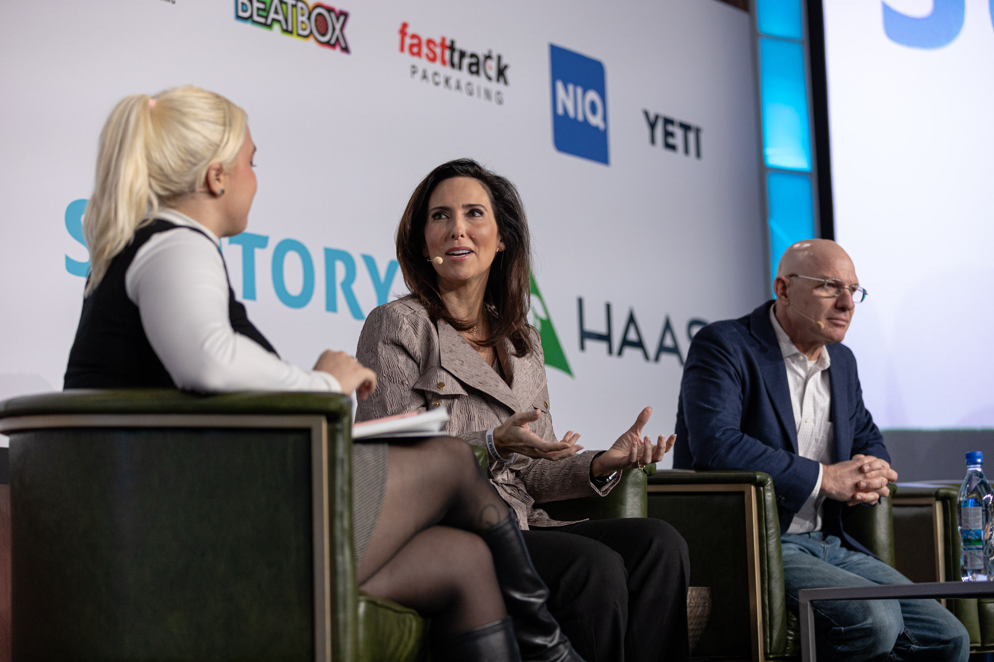 A woman in a blonde ponytail interviews two seated panelists on stage in green armchairs.