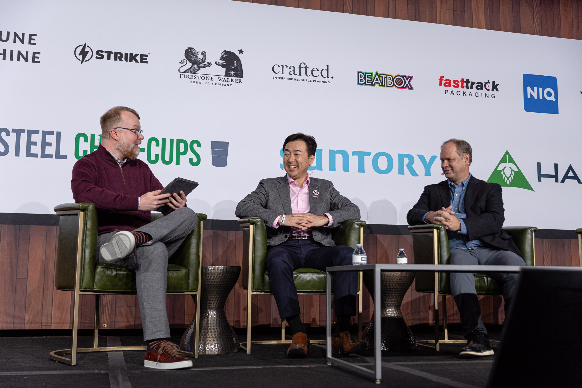 Three men sit in green armchairs on stage with the Suntory logo on the wall behind them.