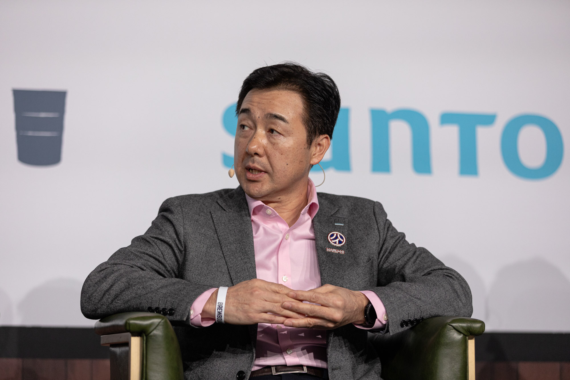 A man in a gray blazer and pink shirt sits in a green armchair in front of a Suntory sign.