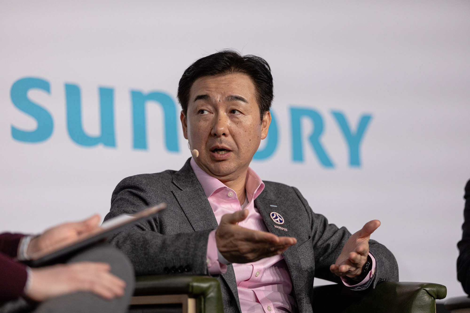 A man in a gray blazer and pink shirt gestures with open hands while seated on stage.