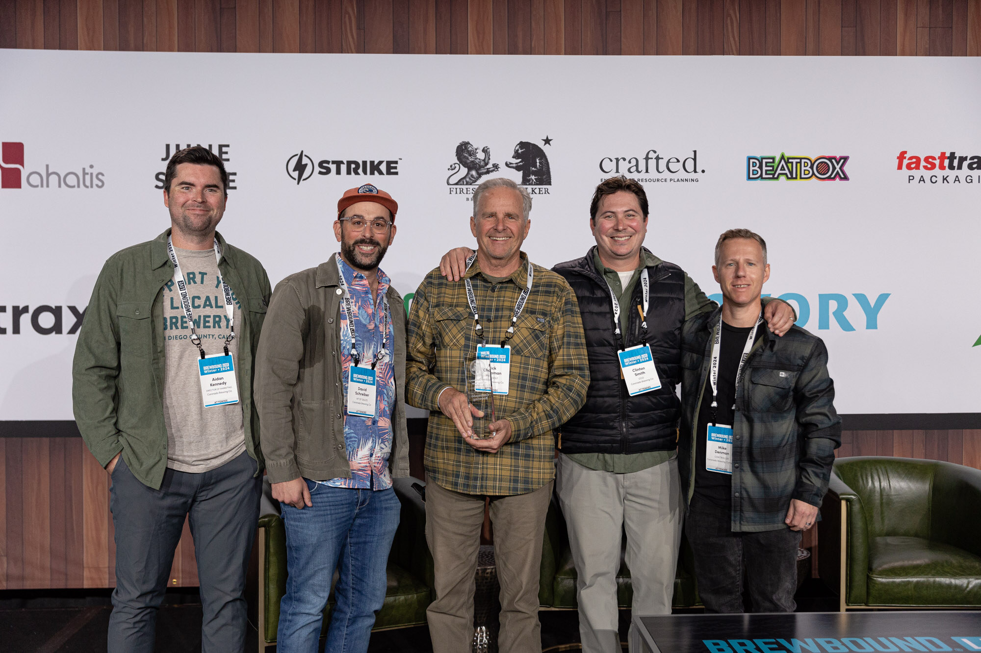 Five men stand shoulder to shoulder on stage, one holding an award, in front of the sponsor wall.