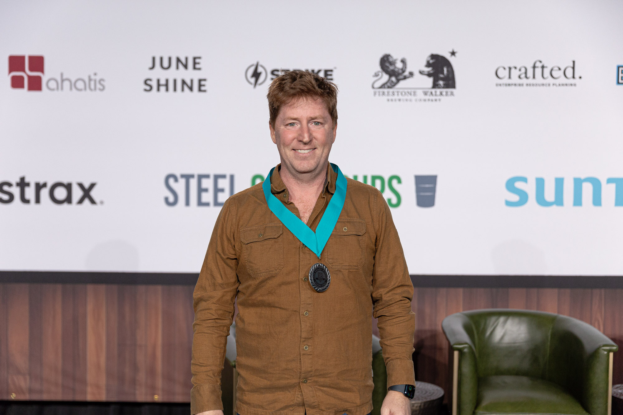 A man in a brown jacket stands on stage wearing a teal ribbon with a medal around his neck.