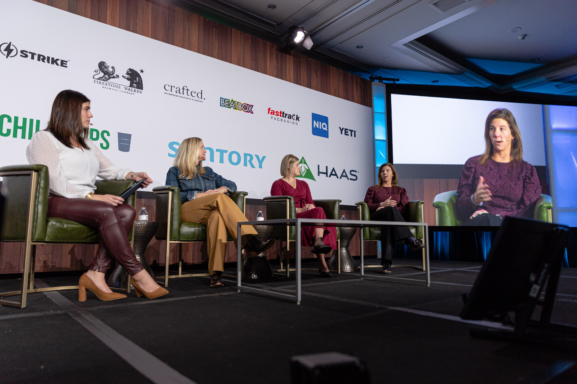 A wide view of five women seated in a row on stage, with one shown larger on a screen at right.