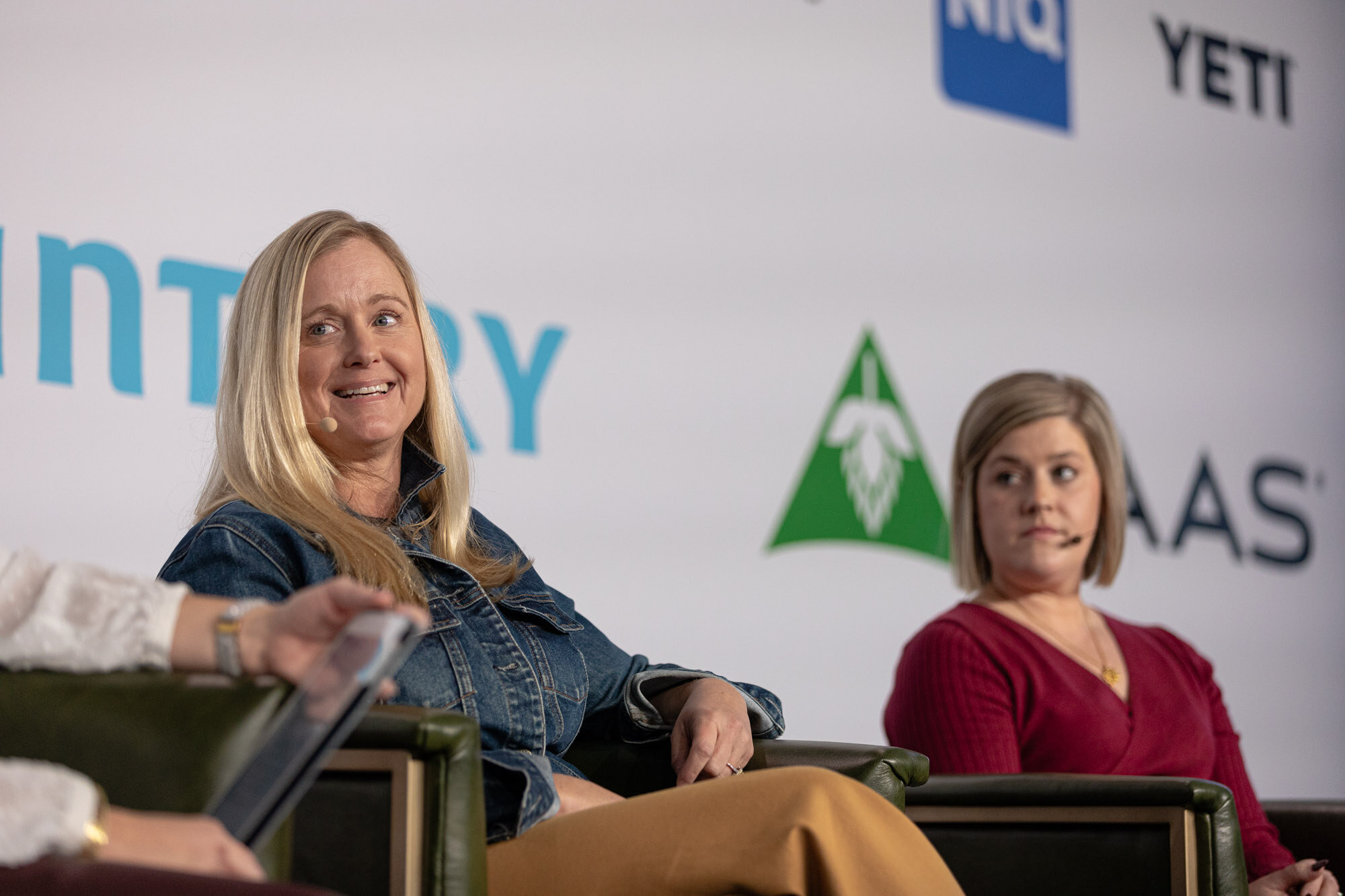 Two women sit in green armchairs on stage, one in a denim jacket and one in a red top.