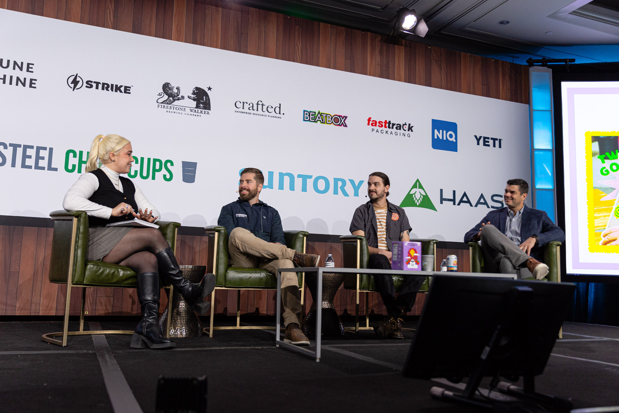 A blonde woman and three men sit in green armchairs on stage with a sponsor wall behind.