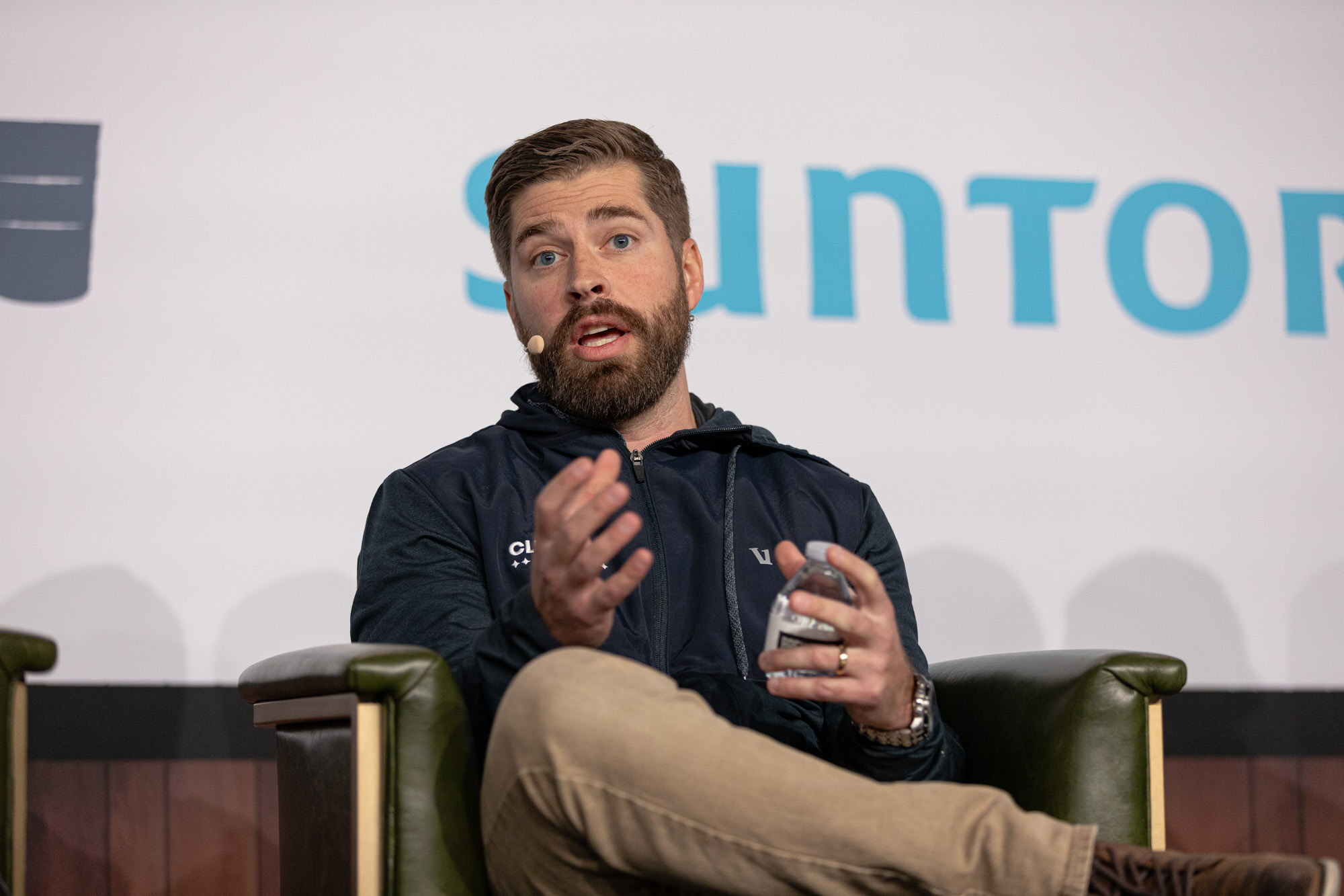 A bearded man in a navy jacket gestures while seated in front of a Suntory banner.