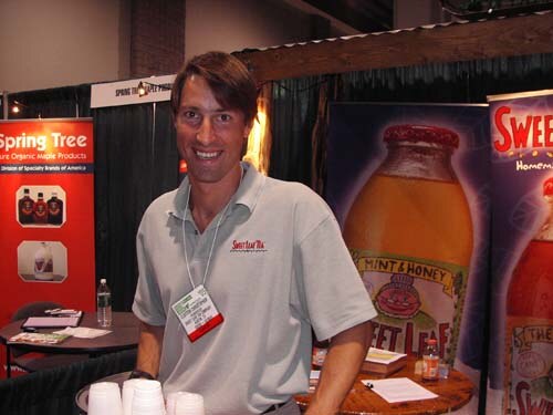 A smiling man in a gray Sweet Leaf Tea polo with a badge stands at a booth. Banners for Spring Tree and a Sweet Leaf Tea Mint & Honey bottle are behind him, with cups on the table.