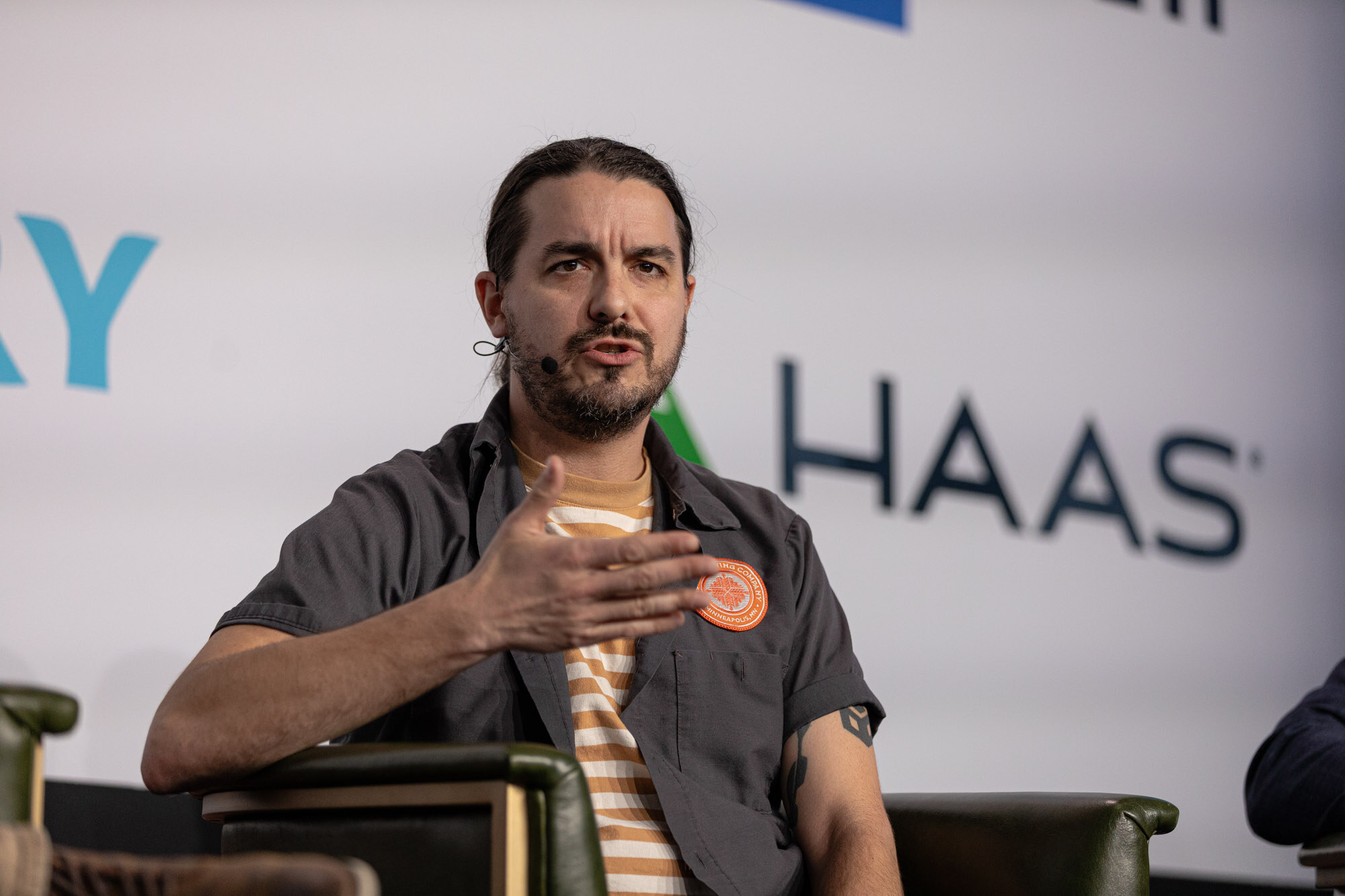 A man with a ponytail and a striped shirt under a gray shirt gestures while seated on stage.