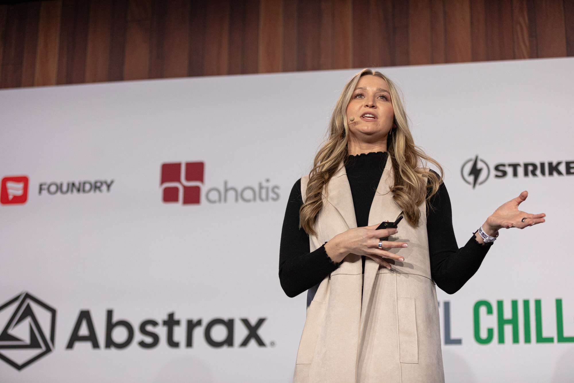 A woman in a cream vest and black top gestures while speaking on stage in front of the sponsor wall.