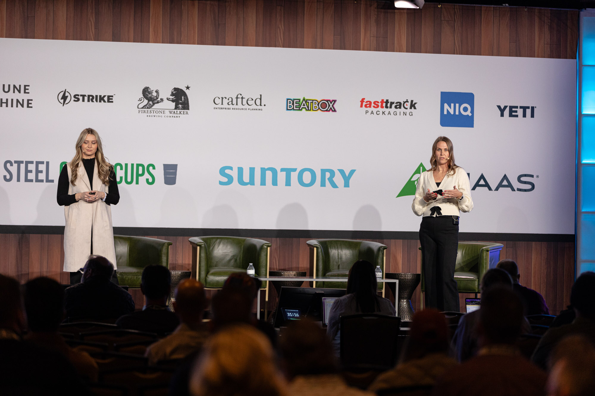 Two women stand on stage in front of green armchairs and an audience with the sponsor wall behind them.