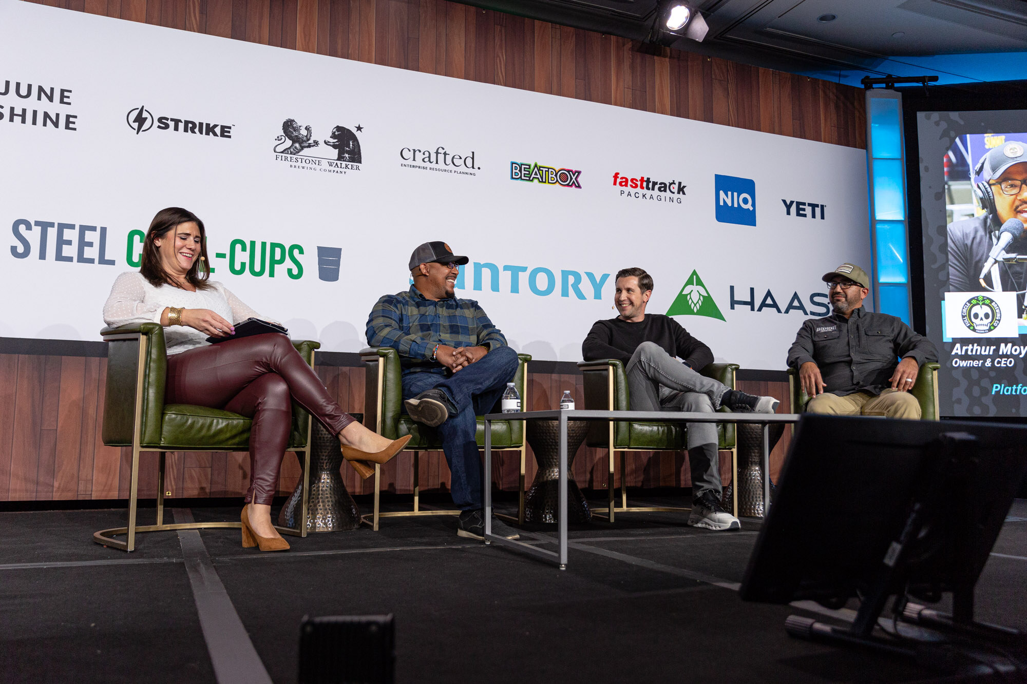 A woman and three men sit on stage while a speaker's name is shown on a screen at the right edge.