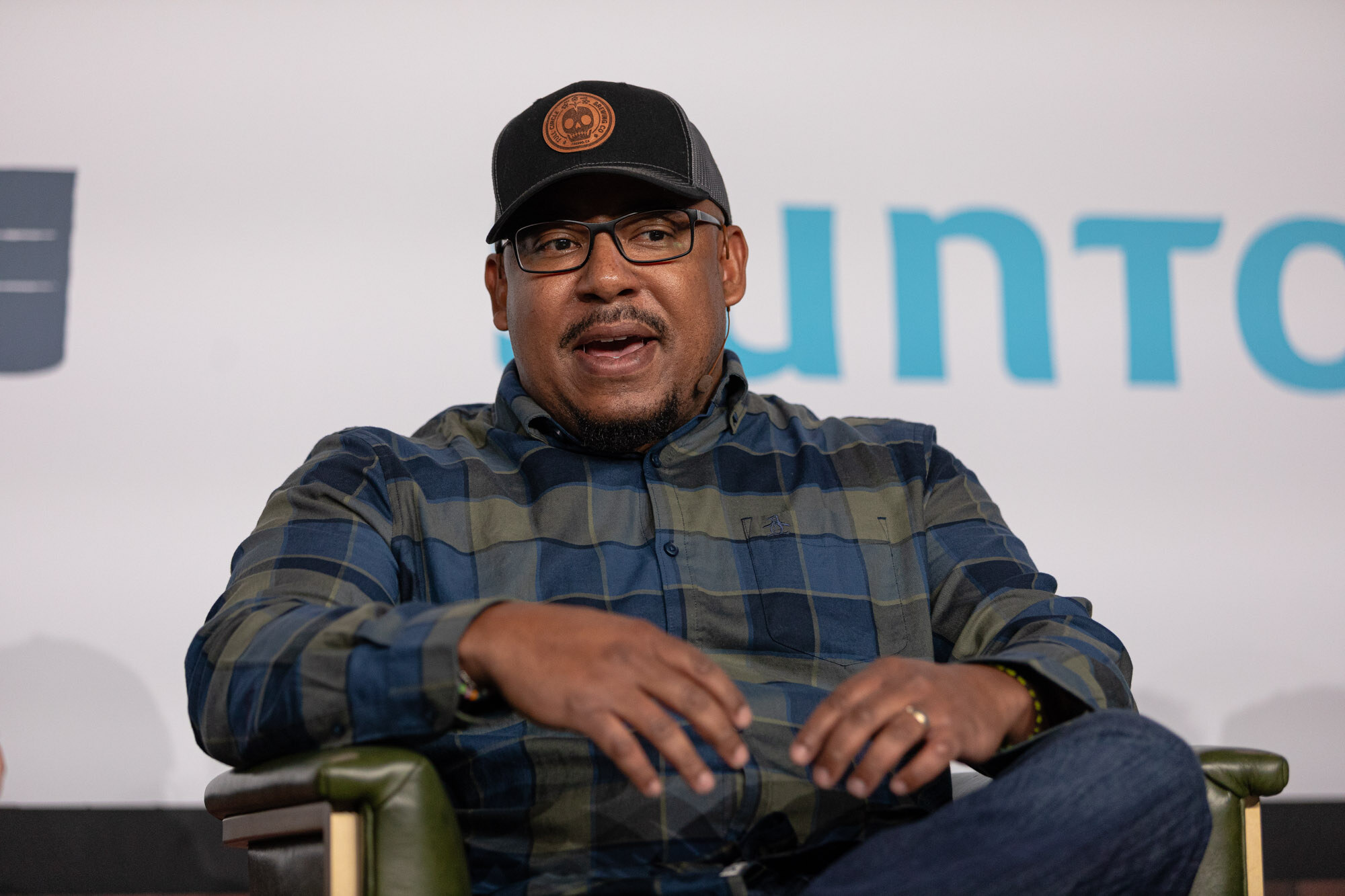 A man in a plaid shirt speaks while seated on stage in front of a Suntory sign.
