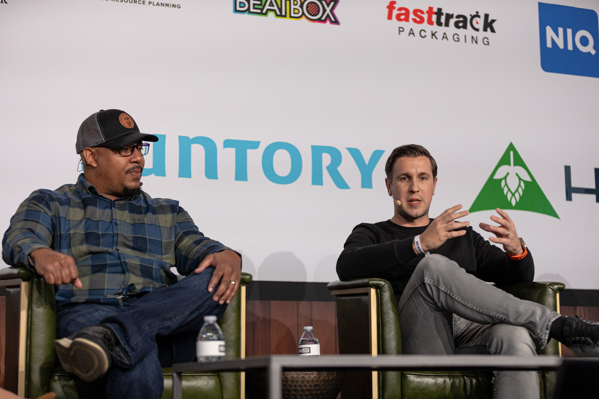Two men sit in green armchairs on stage, one gesturing with both hands.