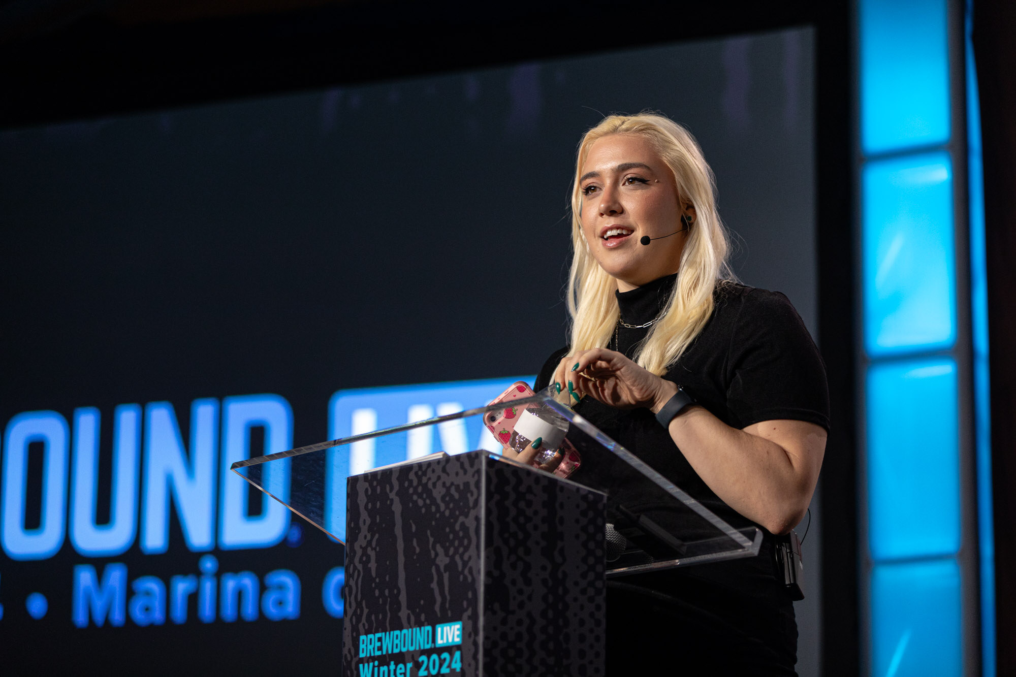 A blonde woman speaks from a podium on a dark stage with a Brewbound Live banner.