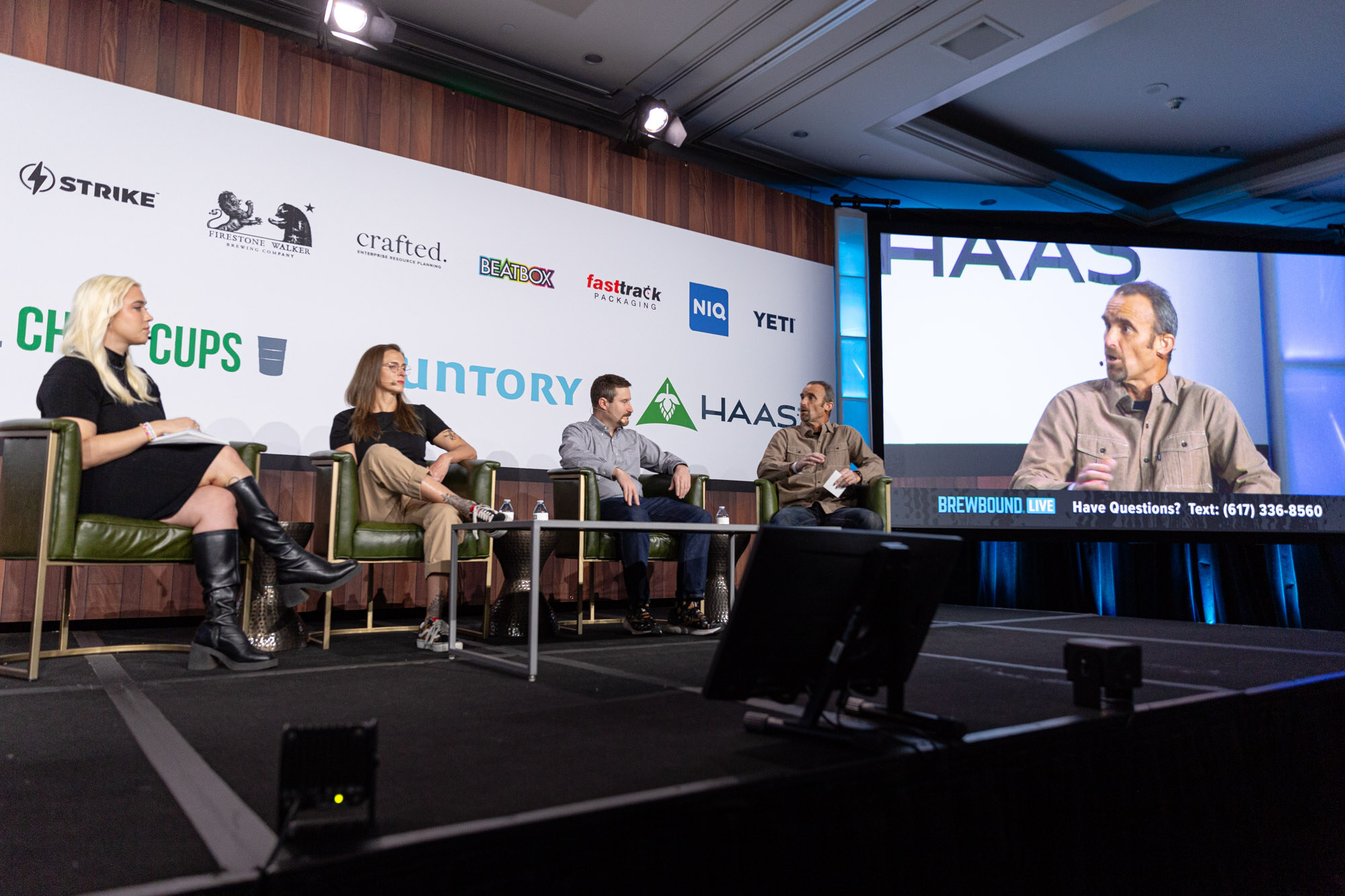 Four people sit on stage in green armchairs while a man appears larger on a screen at right.