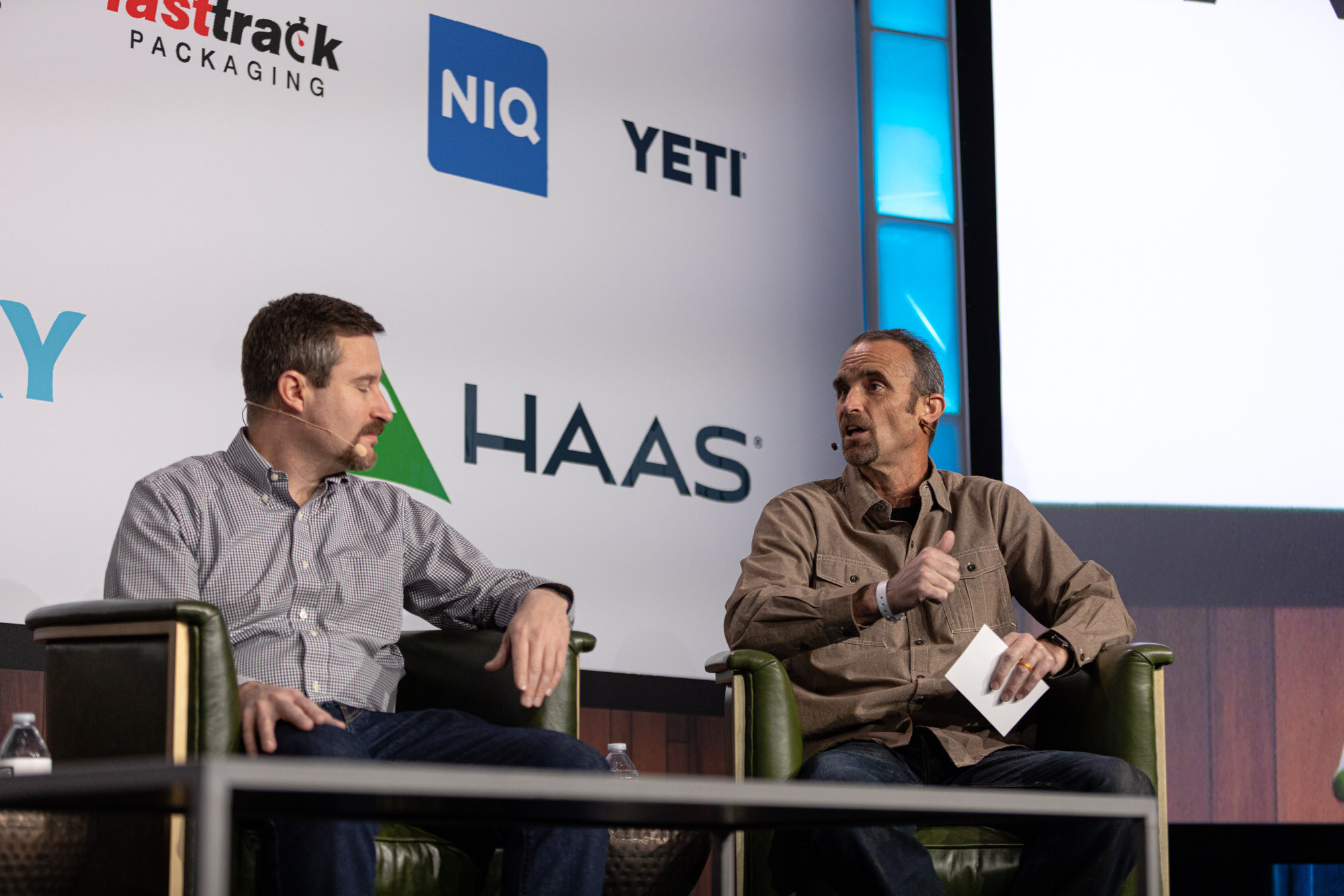 Two men sit on stage in front of a Haas sign, one in a gray shirt and one in a tan shirt holding paper.