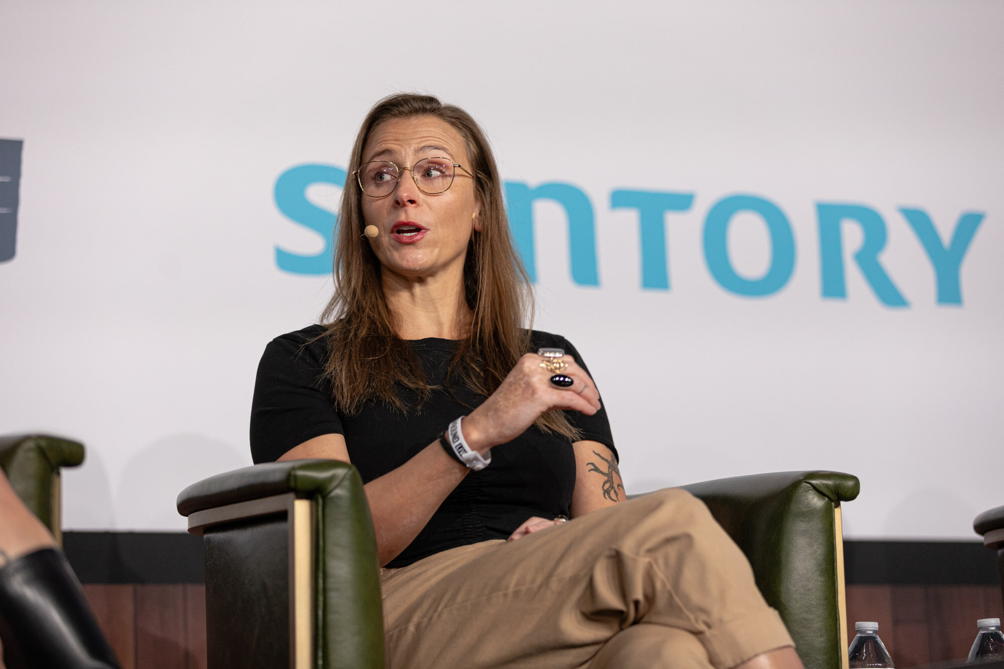 A woman with glasses and a black top speaks while seated in front of a Suntory sign.