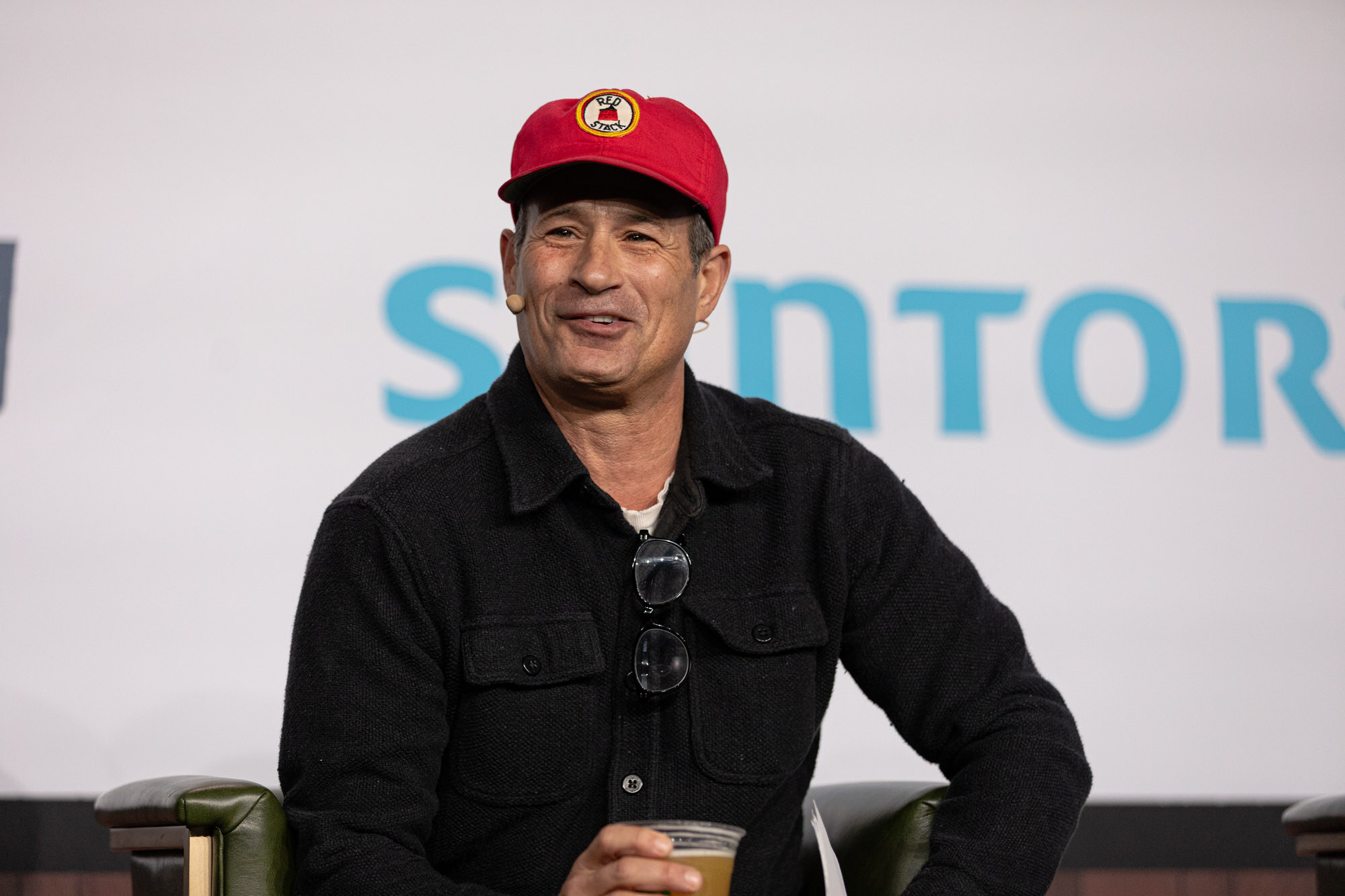 A man in a red cap and black jacket smiles while seated in front of a Suntory sign.
