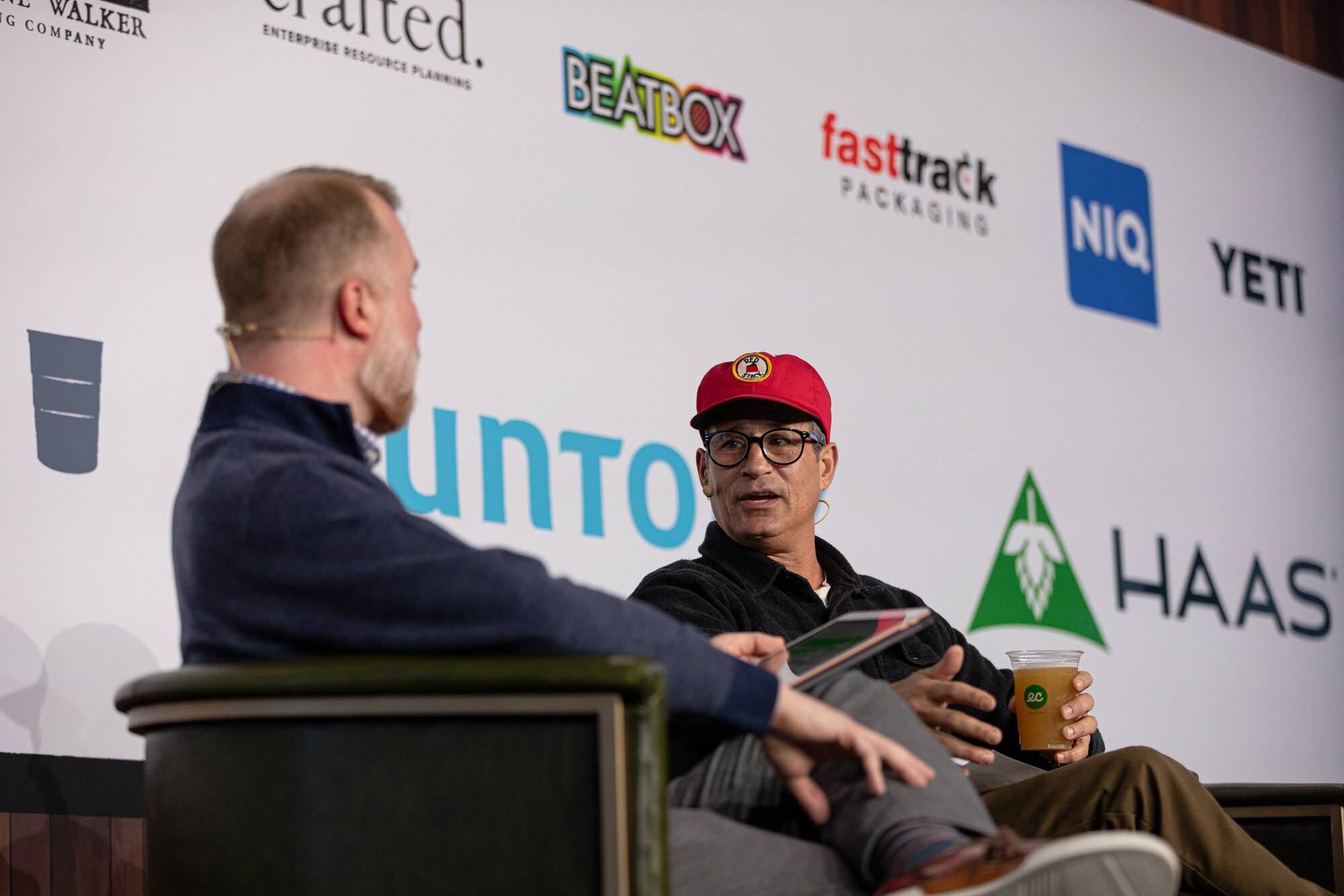 Two men sit side by side on stage, one in a red cap holding a drink.