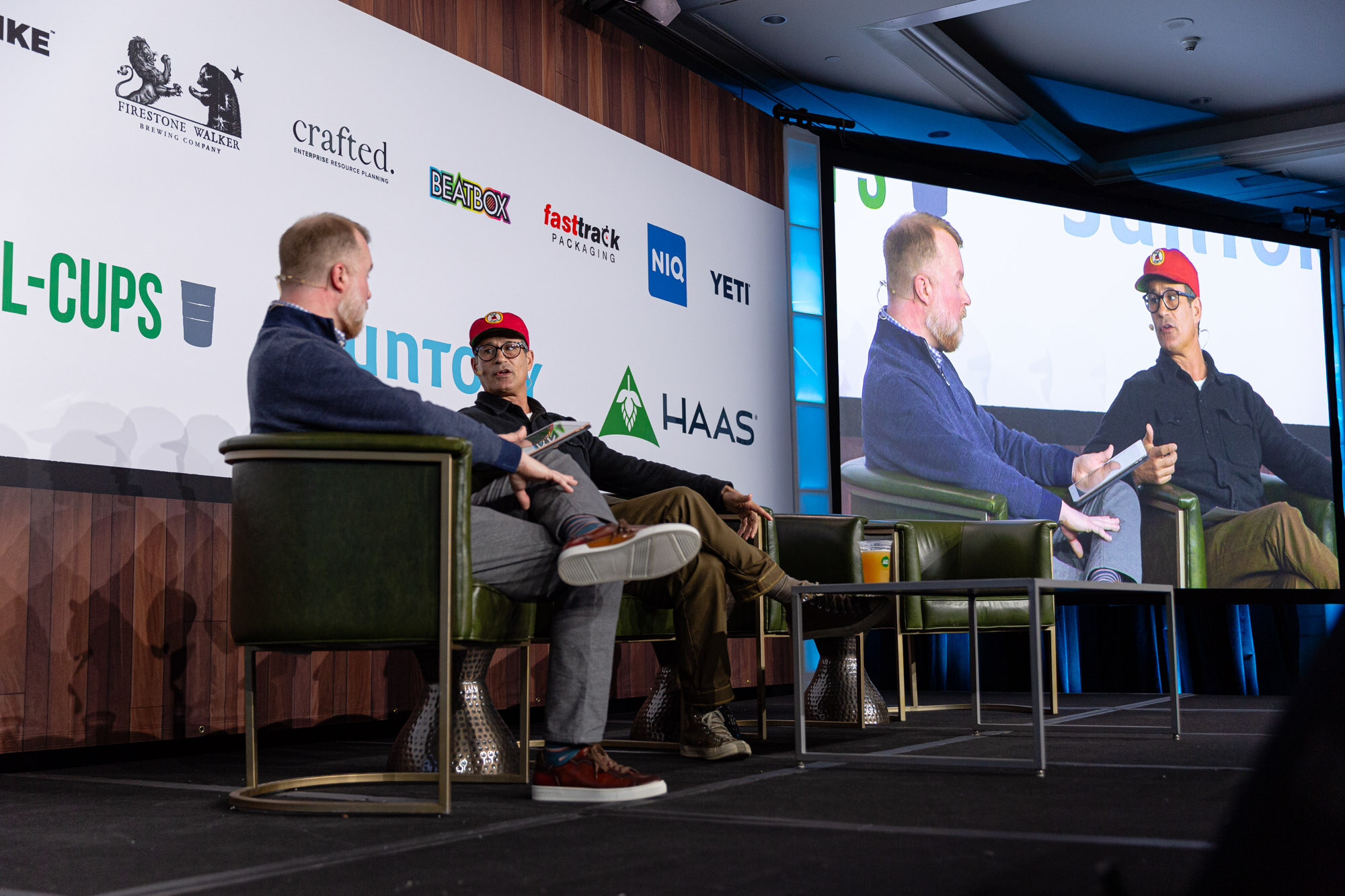 Two men sit in green armchairs, one in a red cap, with a large screen behind them.