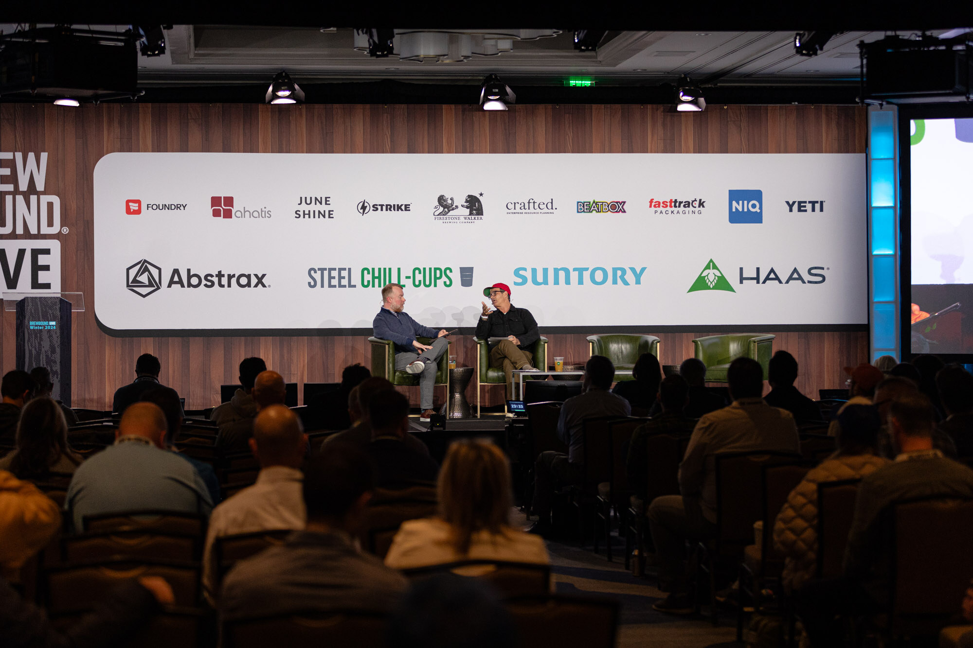 Two men sit in armchairs on a stage in front of a seated audience and the sponsor wall.