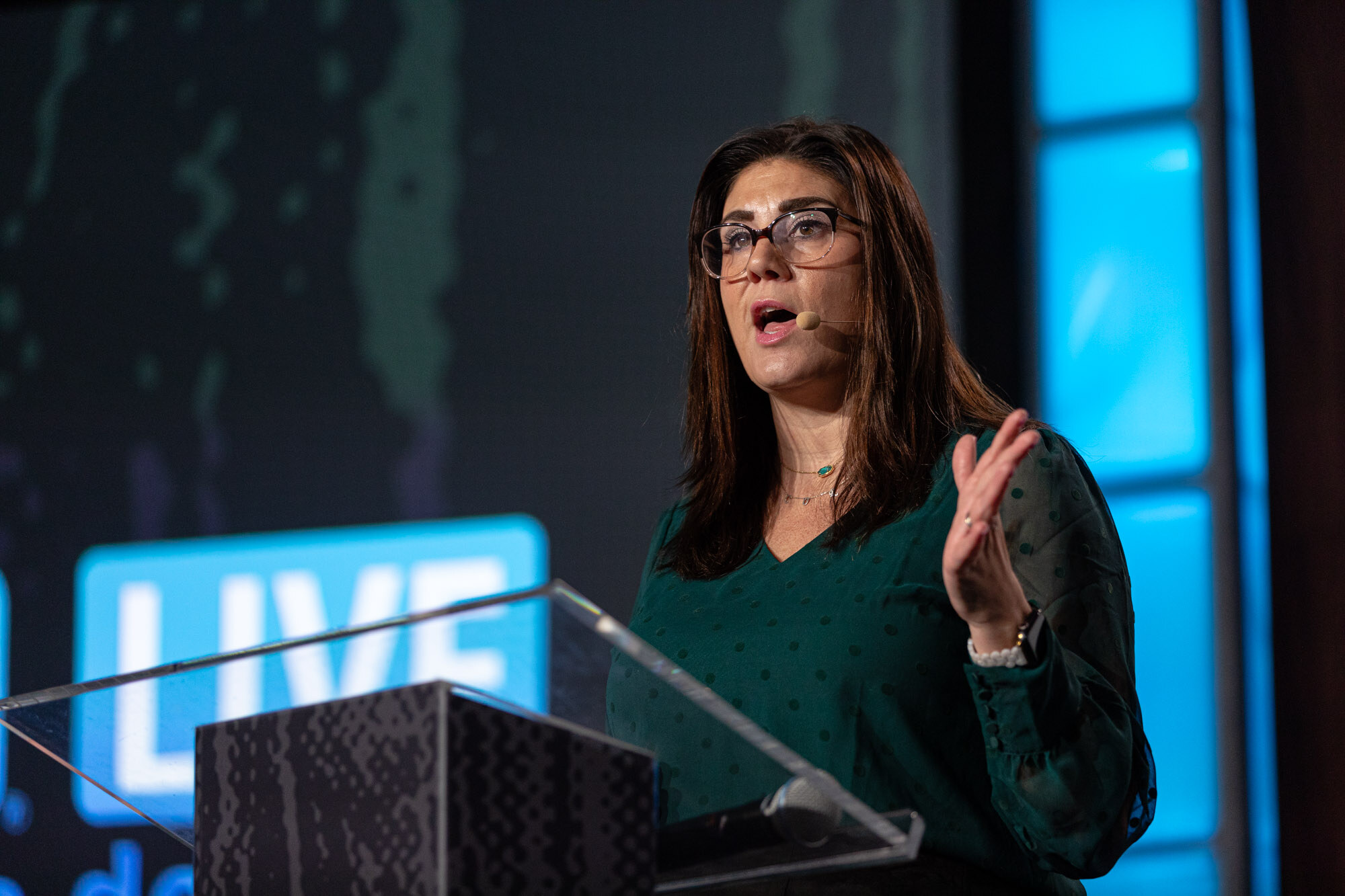 A woman in a green blouse speaks from a podium on stage.