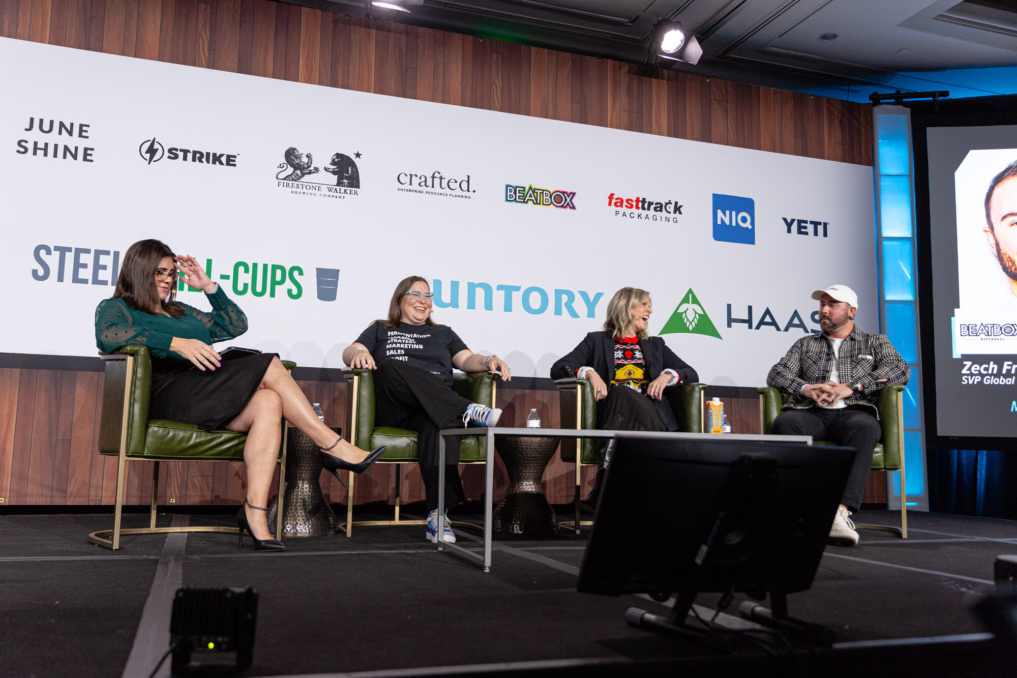 Four people sit in a row on stage with a speaker's photo shown on a screen at the right.