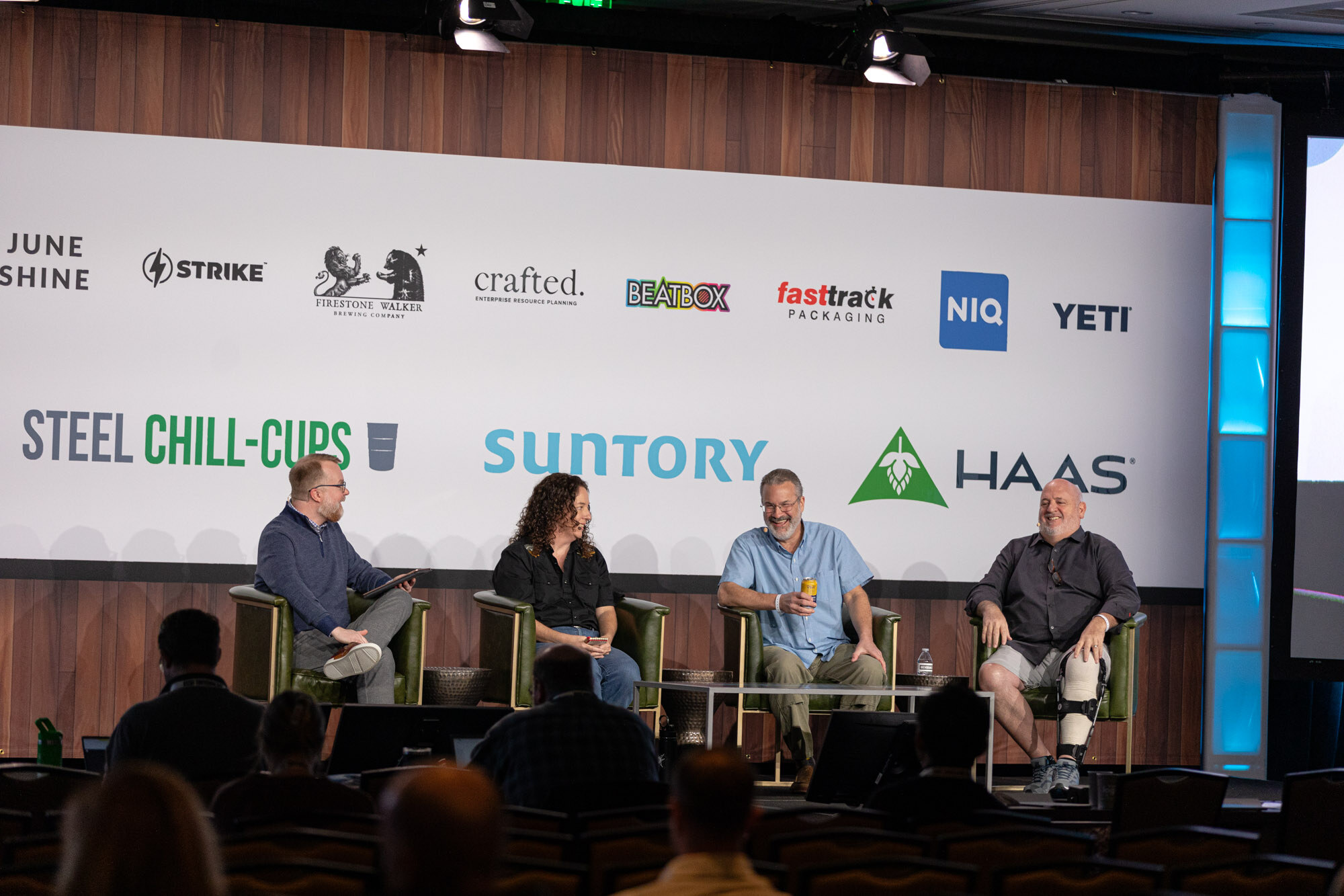 Four people sit in green armchairs in front of the sponsor wall with some audience heads in the foreground.