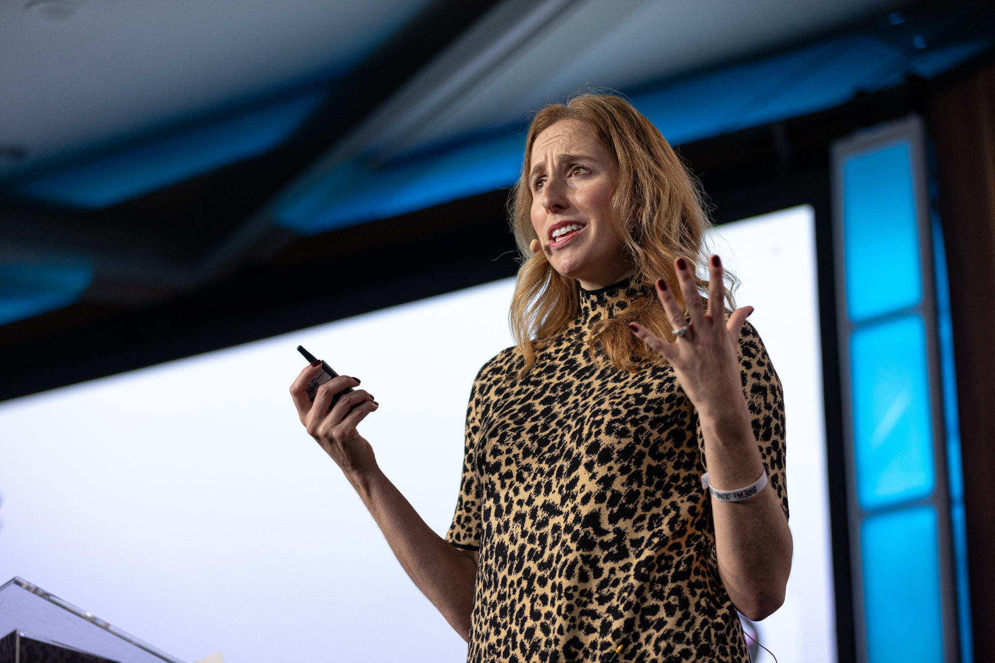 A woman in a leopard-print dress holds a remote and gestures on stage.