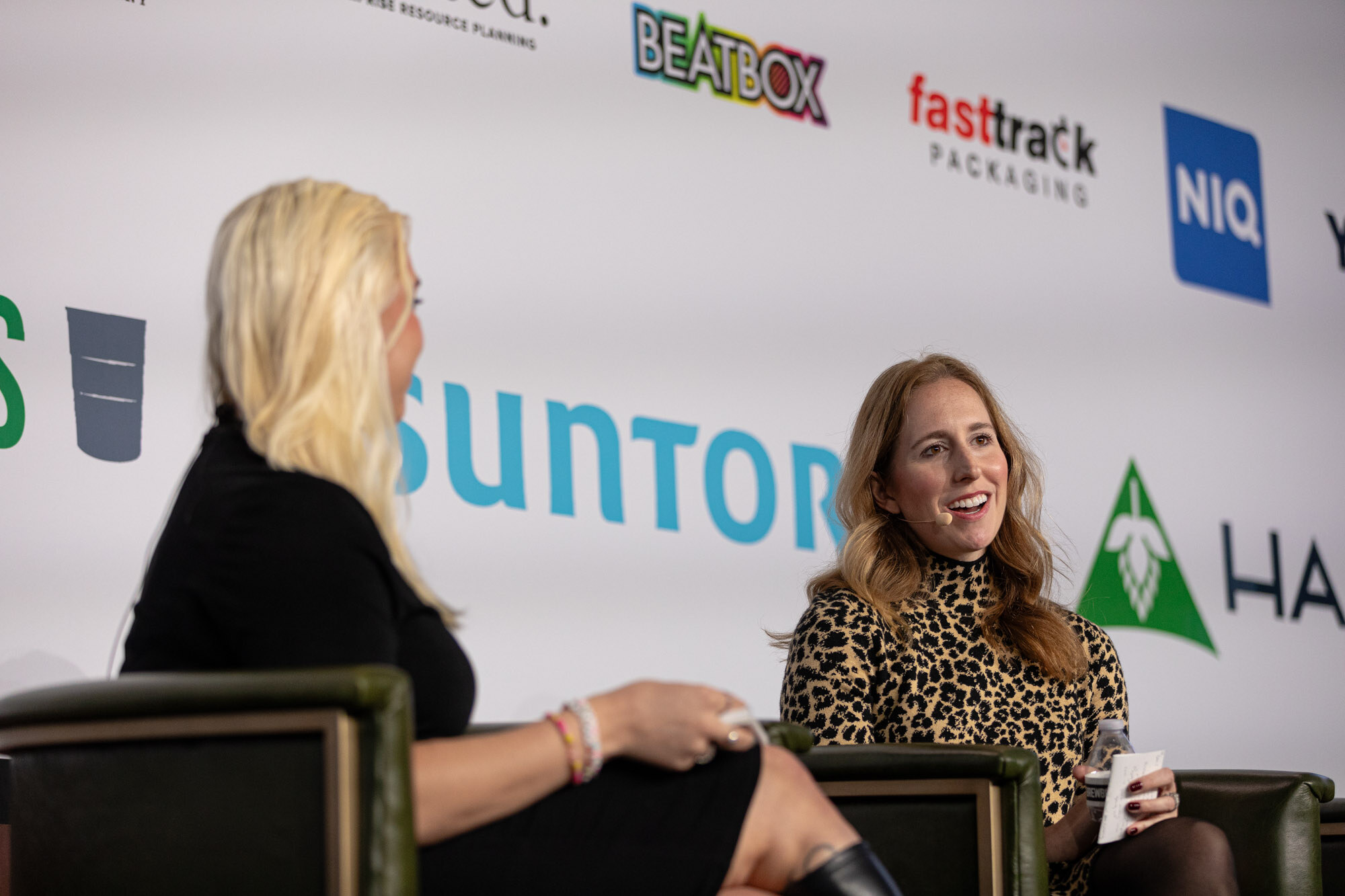 Two women sit on stage, one in a leopard-print dress smiling at the other.