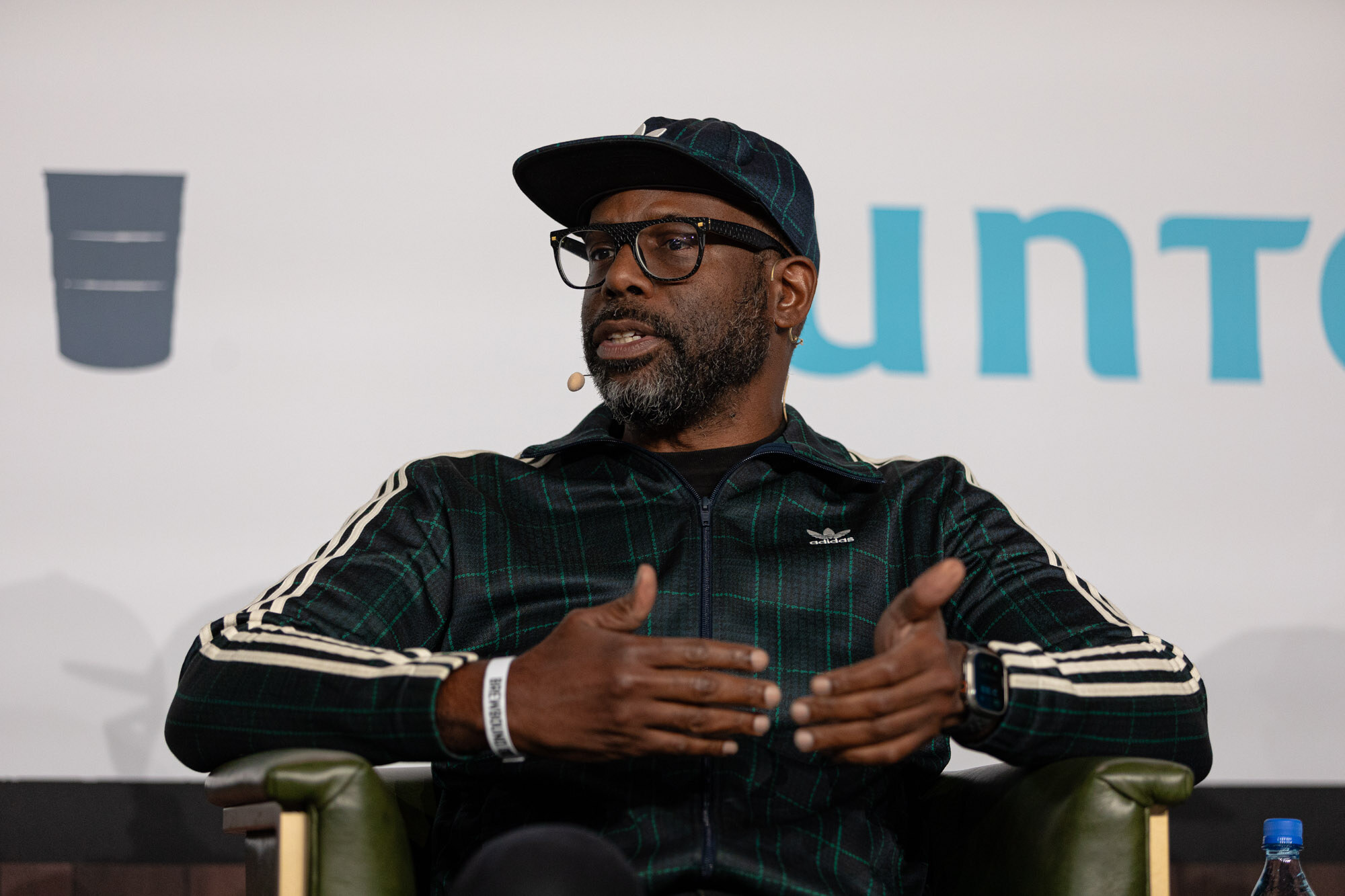 A man in a dark green striped jacket and cap sits in an armchair and speaks.