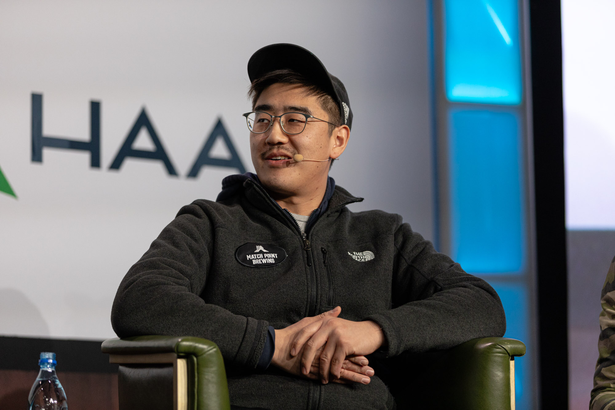 A man in a black cap and glasses sits in front of a Haas sign.