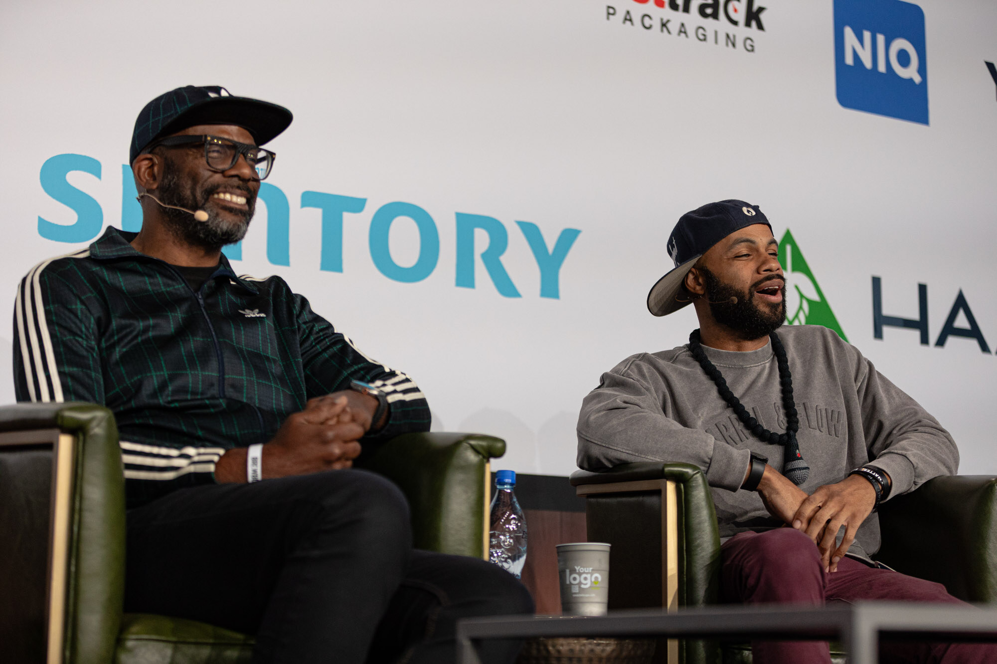 Two men in caps sit in armchairs on stage, one in a green striped jacket, both laughing.