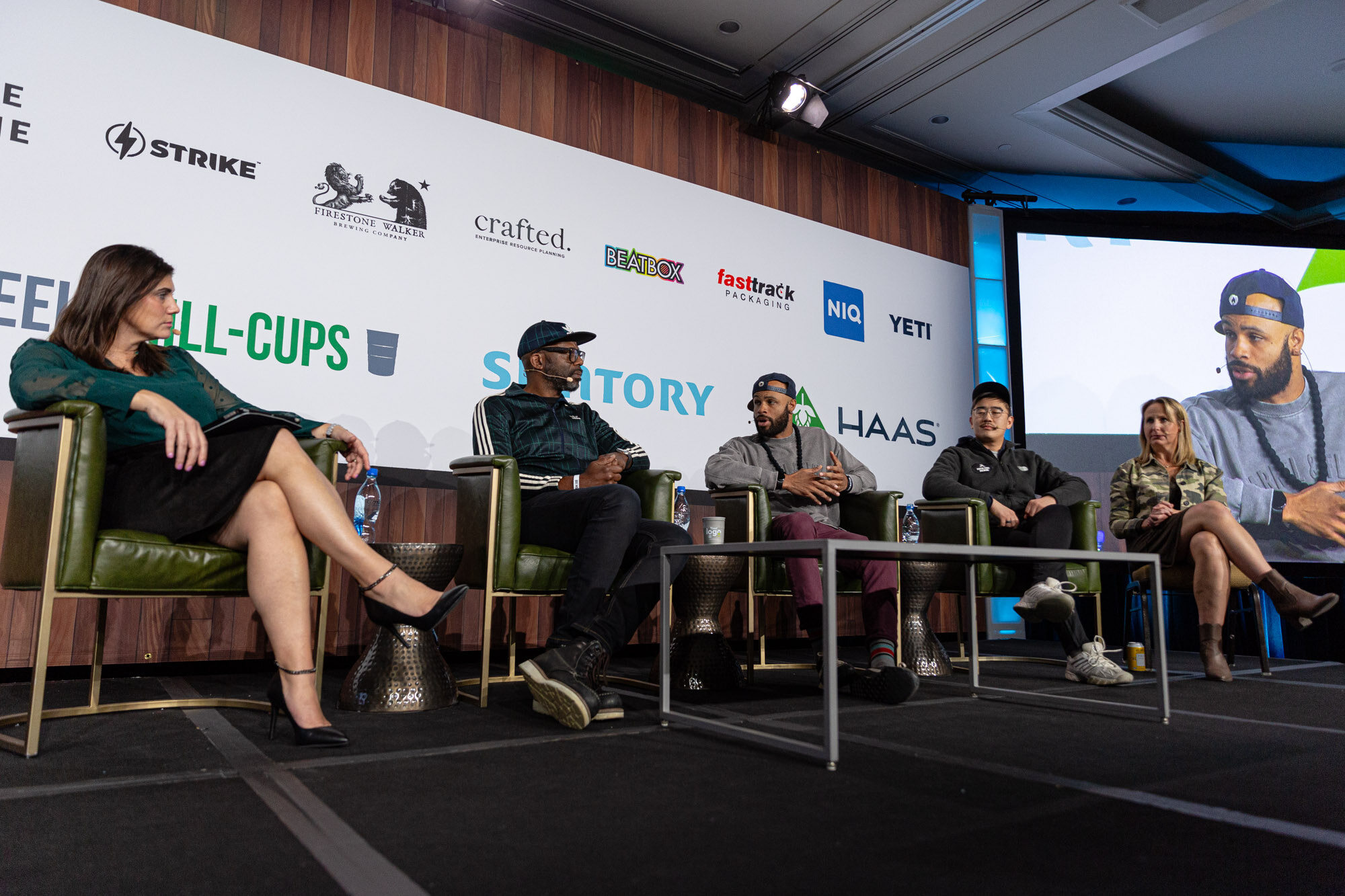 A woman and four men sit in a row on stage in front of the sponsor wall with a screen at right.