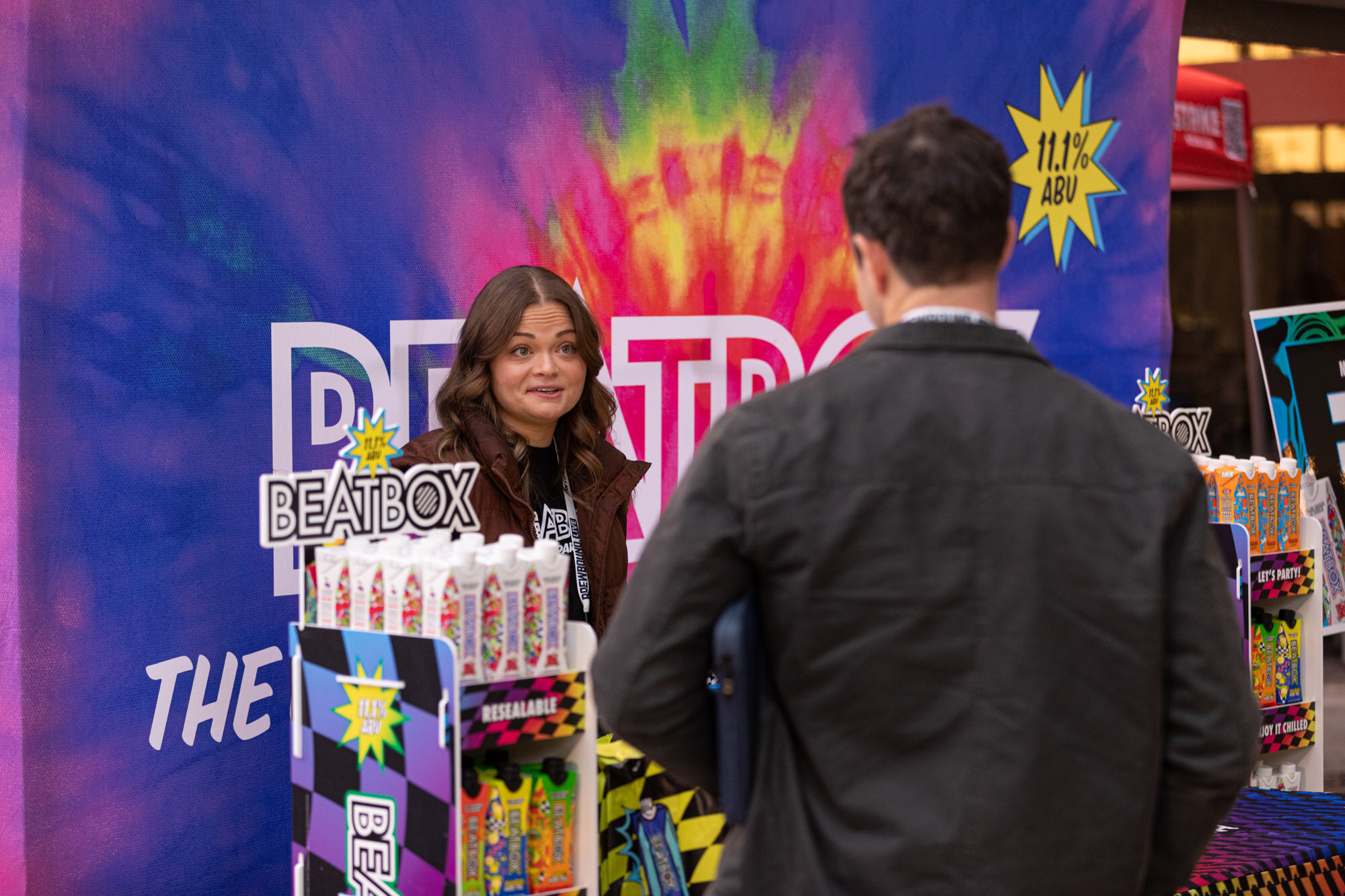 A woman stands in front of a colorful Beatbox backdrop talking to a man in a black jacket.