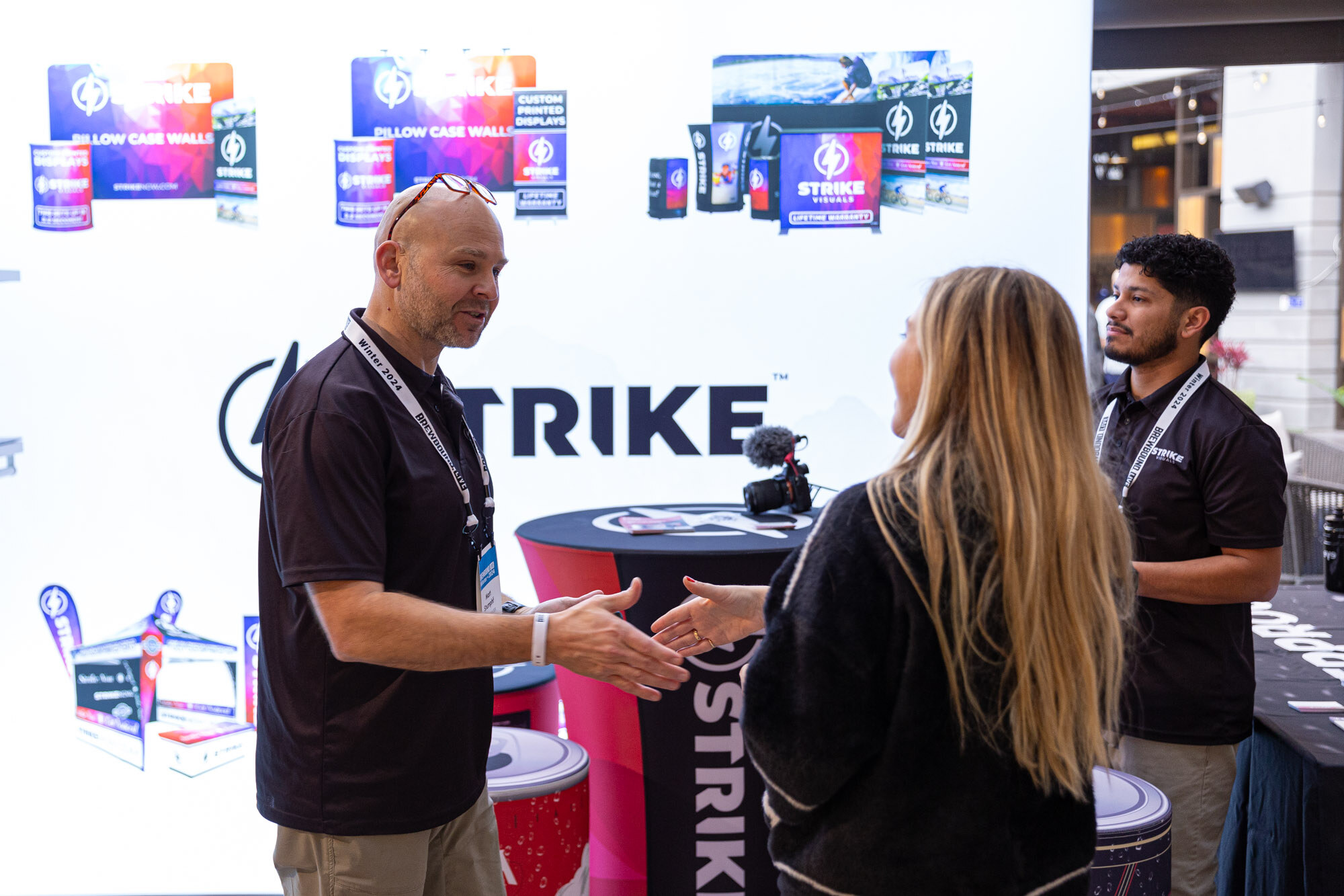 A man in a black polo shakes hands with a woman at a Strike booth.