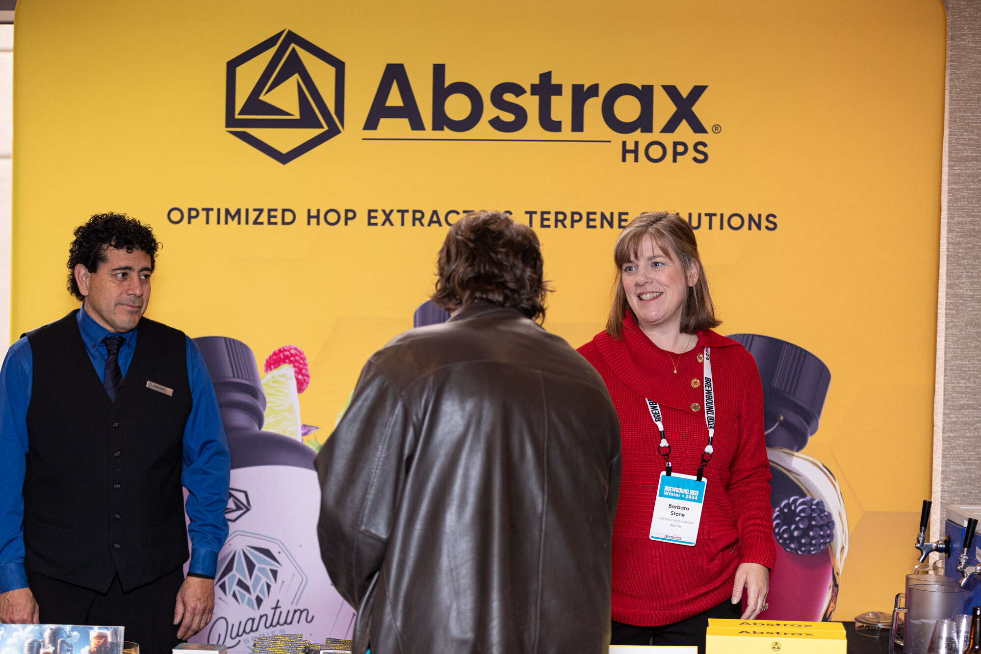 Two Abstrax staff stand in front of a yellow Abstrax Hops banner talking with an attendee.