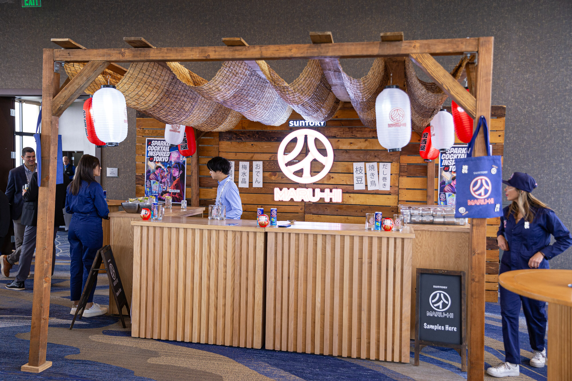 A wooden bar with red paper lanterns and a Maru-Hi sign, with staff in blue coveralls.