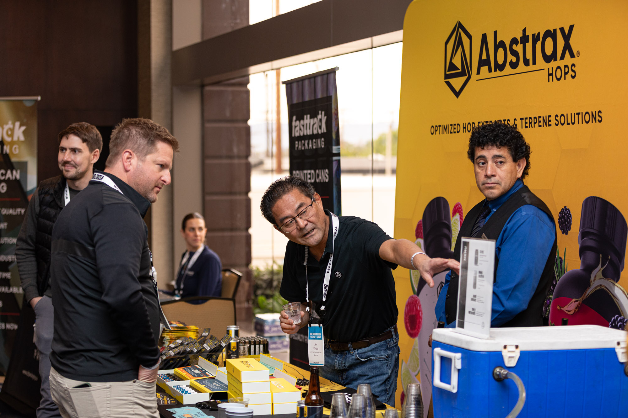Two men stand at an Abstrax Hops booth with a blue cooler and a Fasttrack Packaging banner.