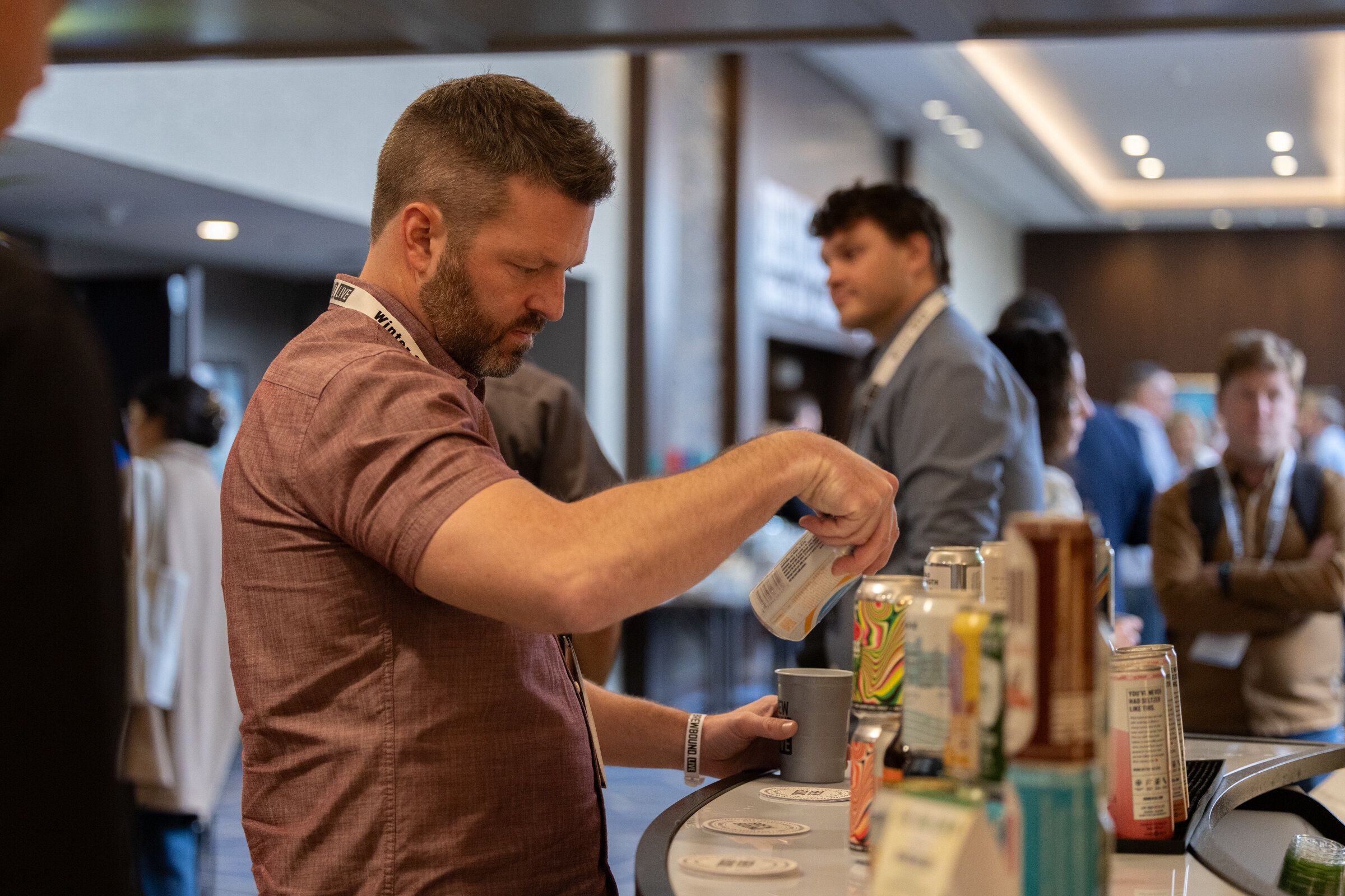 A man in a rust-colored shirt pours a can into a cup at a bar lined with cans.