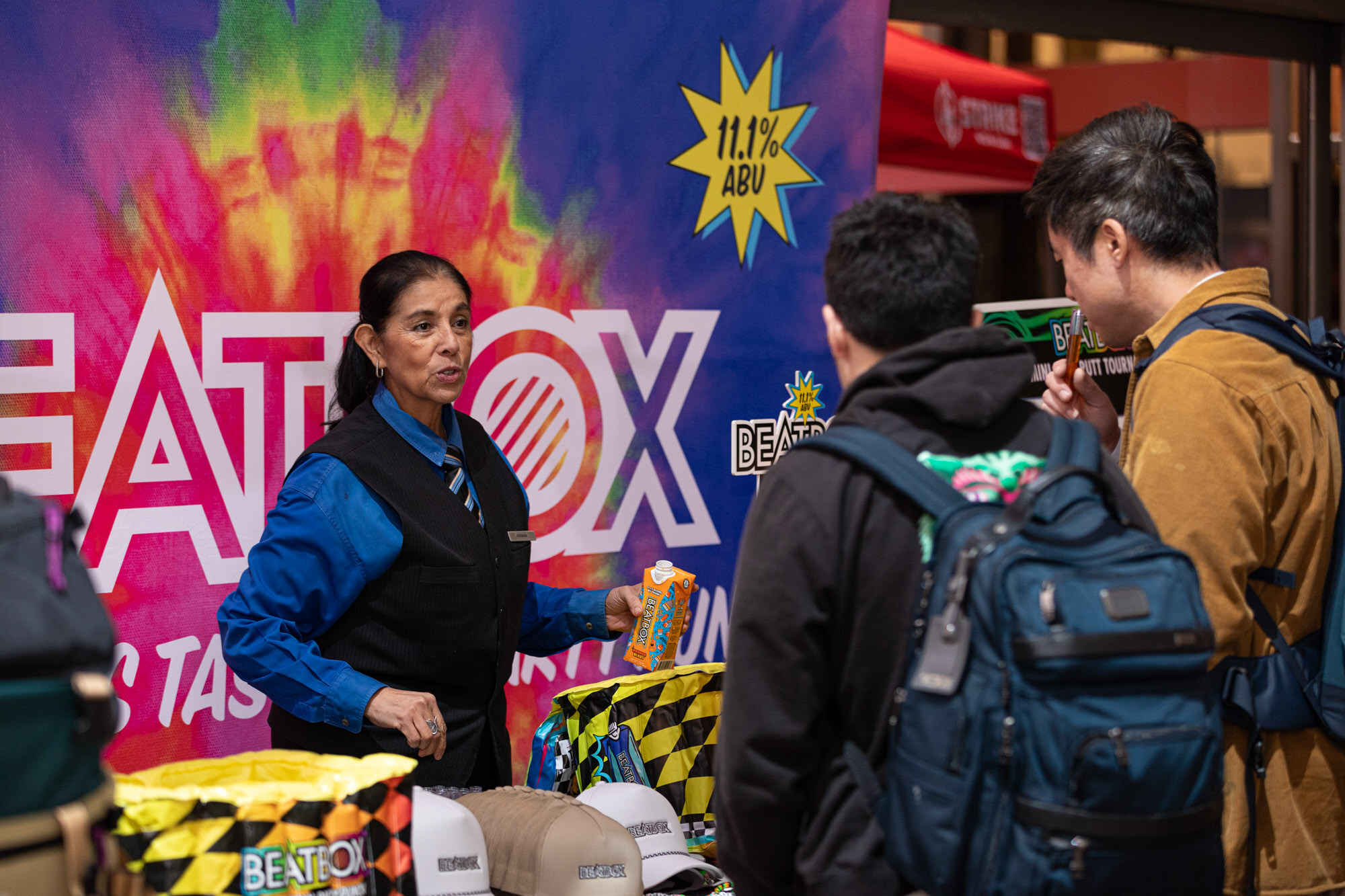 A woman in a blue vest stands in front of a Beatbox backdrop as attendees pass.