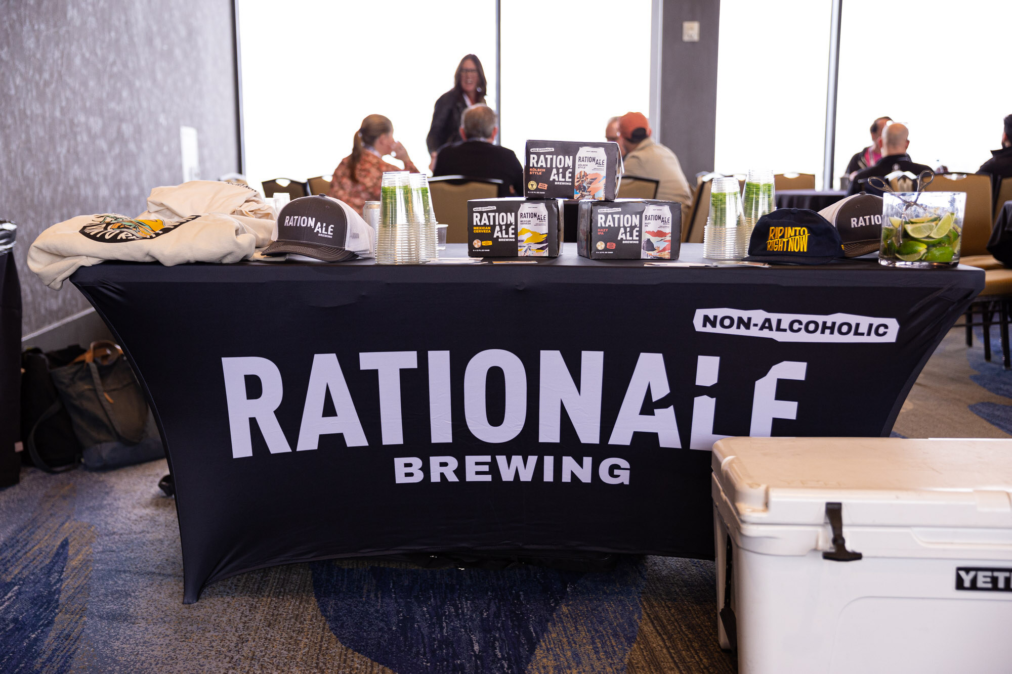 A black table cover reading Rationale Brewing with cans and caps and attendees seated behind it.