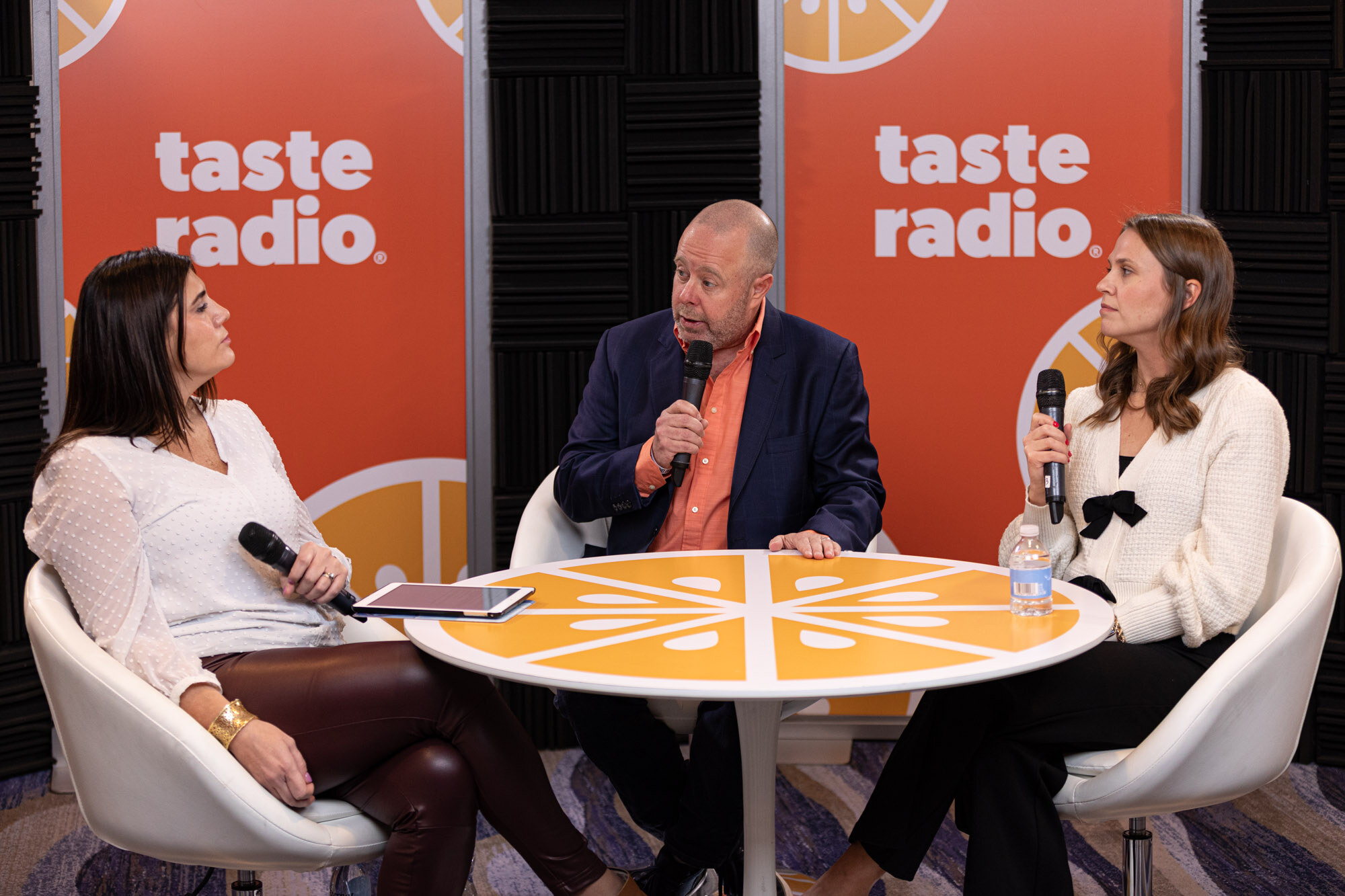Two women and a bald man sit at an orange round table in front of Taste Radio banners.