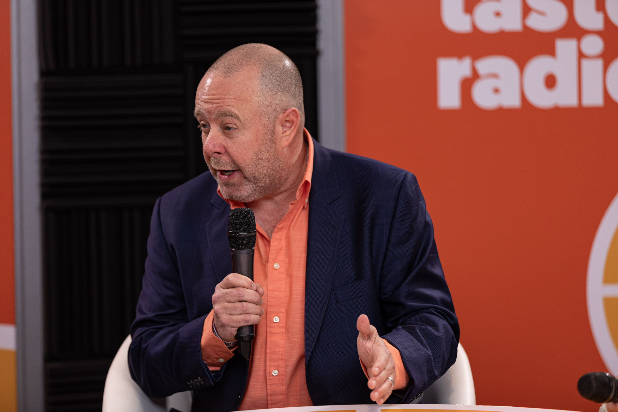 A bald man in a navy blazer and orange shirt speaks into a microphone in front of a Taste Radio banner.