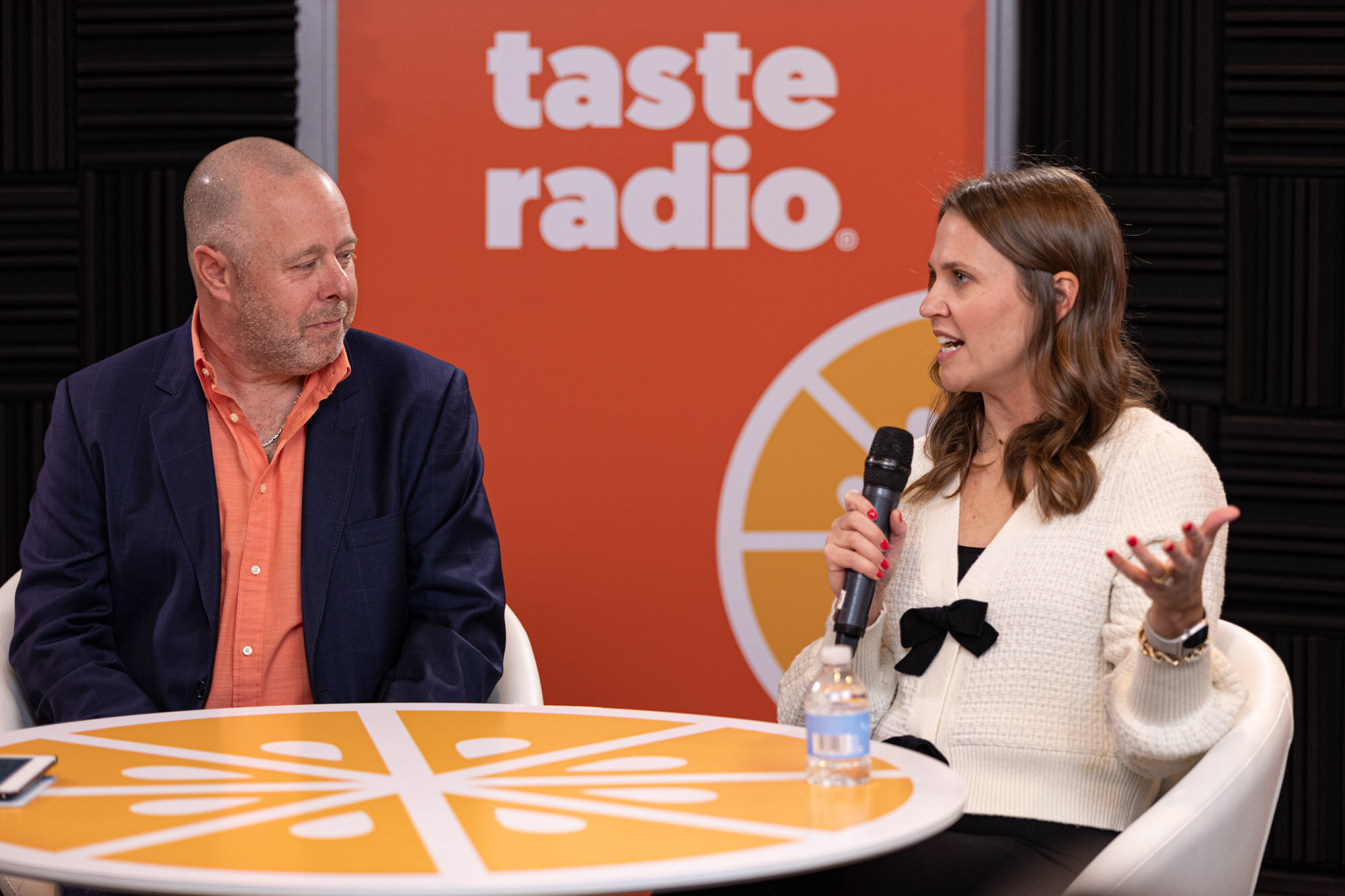 A woman in a cream cardigan speaks into a microphone next to a man at the livestream table.