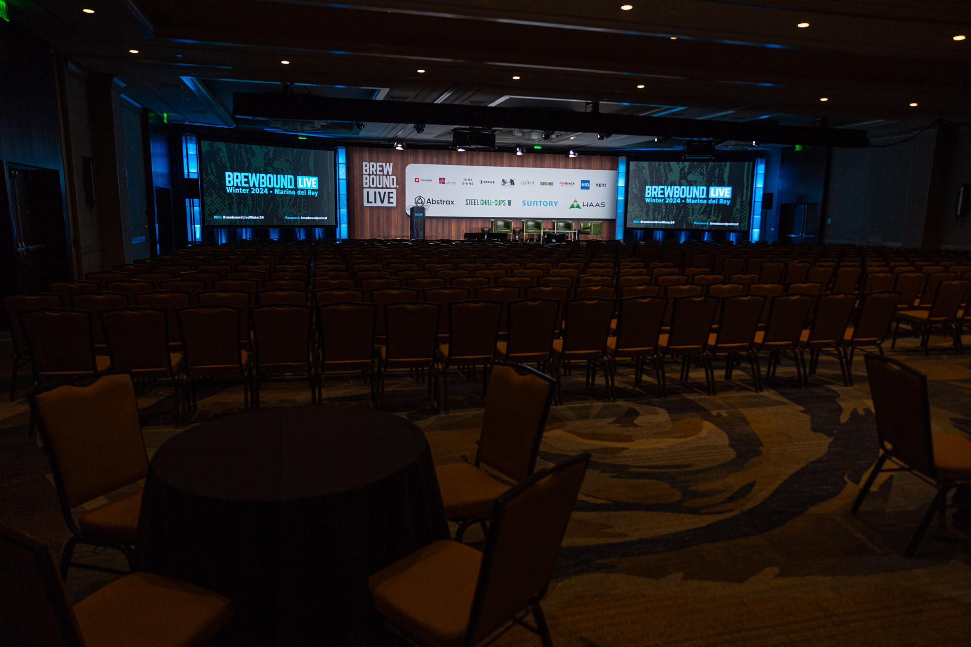 Rows of chairs face a stage with sponsor-wall screens in an empty ballroom.