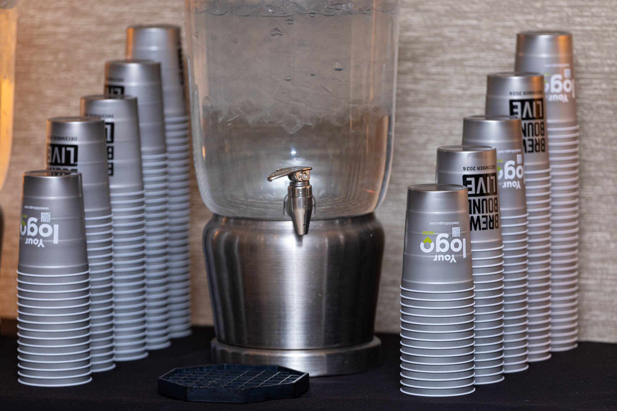 Stacks of gray cups printed with Brewbound Live beside a metal water dispenser.